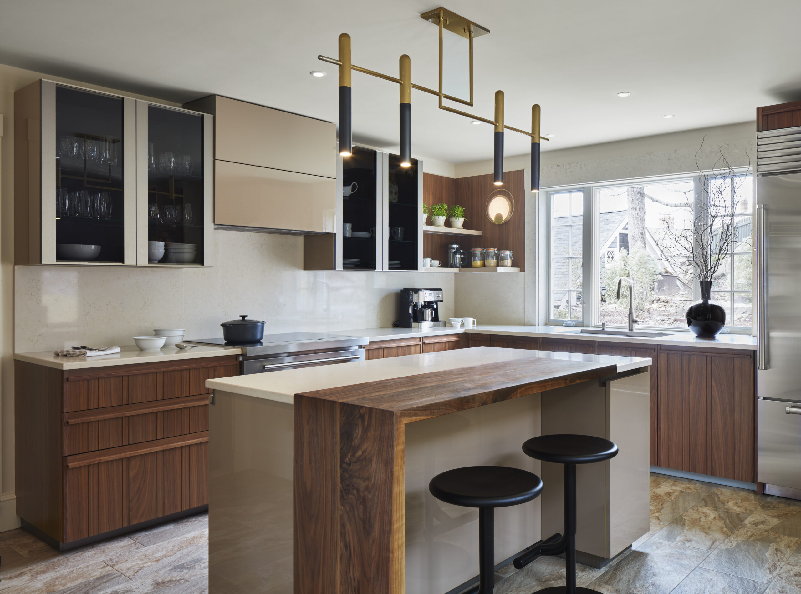 Modern kitchen with wood and beige cabinets, a central island with wooden countertop, two black stools, pendant lighting, and stainless steel appliances near a large window—showcasing luxury interior design by an expert interior designer in Massachusetts.