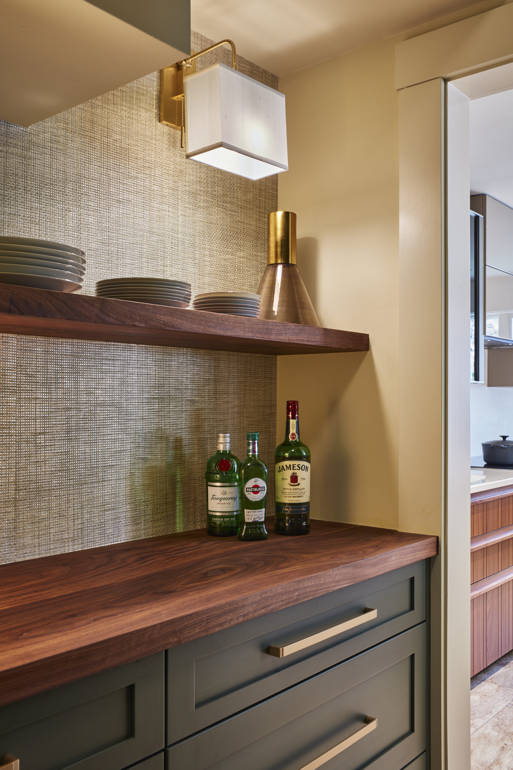 A kitchen bar area with wooden shelves, stacked plates, three bottles of alcohol, a gold vase, and a wall-mounted light fixture above a wooden countertop showcases the elegance of luxury interior design.