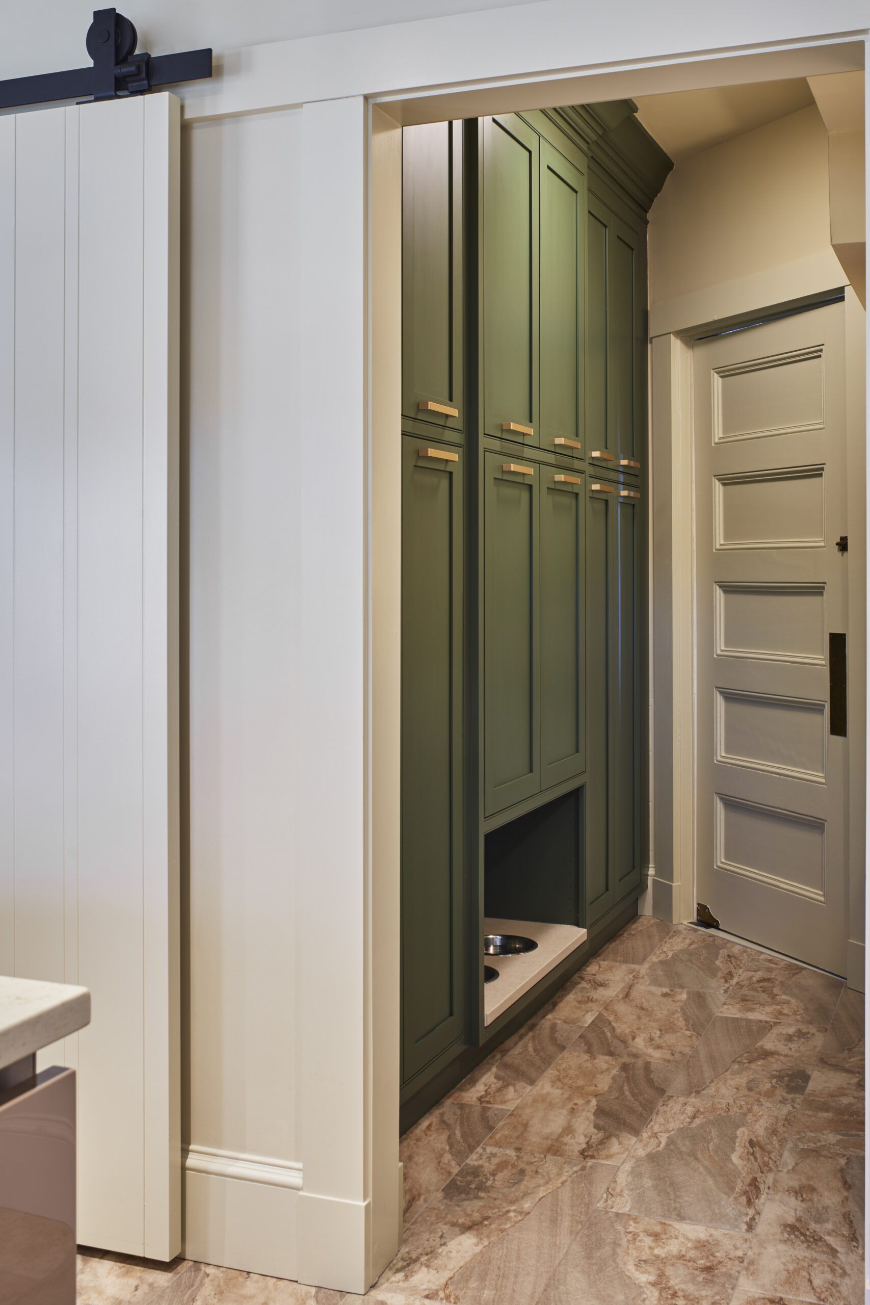 Green built-in cabinets with gold handles bring Luxury Interior Design to this hallway, featuring a recessed area with a pet food bowl on a marble-patterned floor.