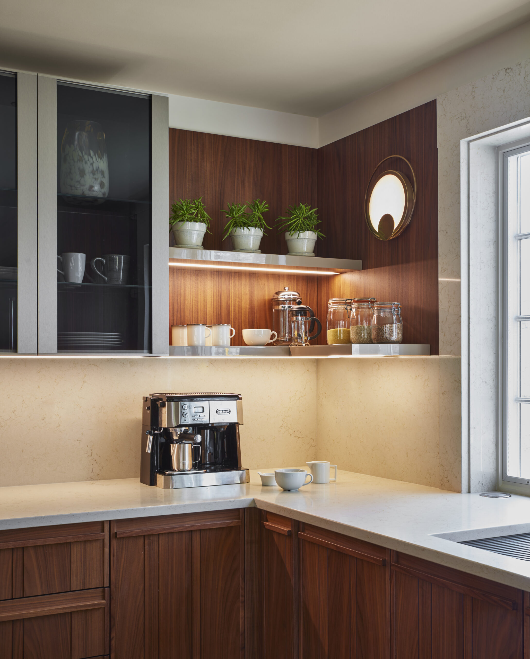 Modern kitchen corner by a top Interior Designer Massachusetts, featuring a coffee maker, French press, jars, potted plants, a round wall light, and ample natural light for a touch of luxury interior design.