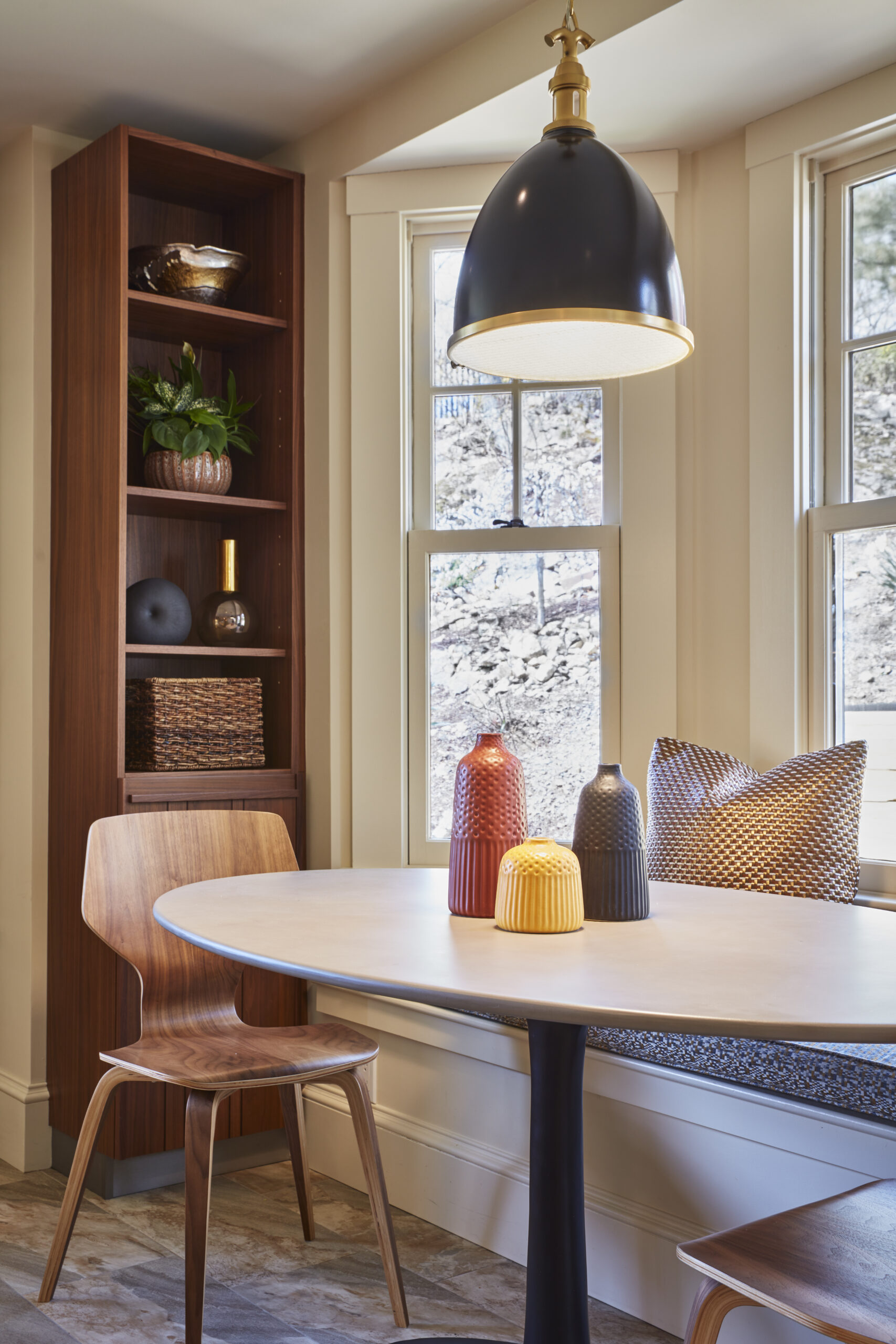 A modern dining nook with a round table, wooden chair, built-in bench with cushions, decorative vases, shelving unit, and a large black pendant light by two windows—an inviting space inspired by luxury interior design.