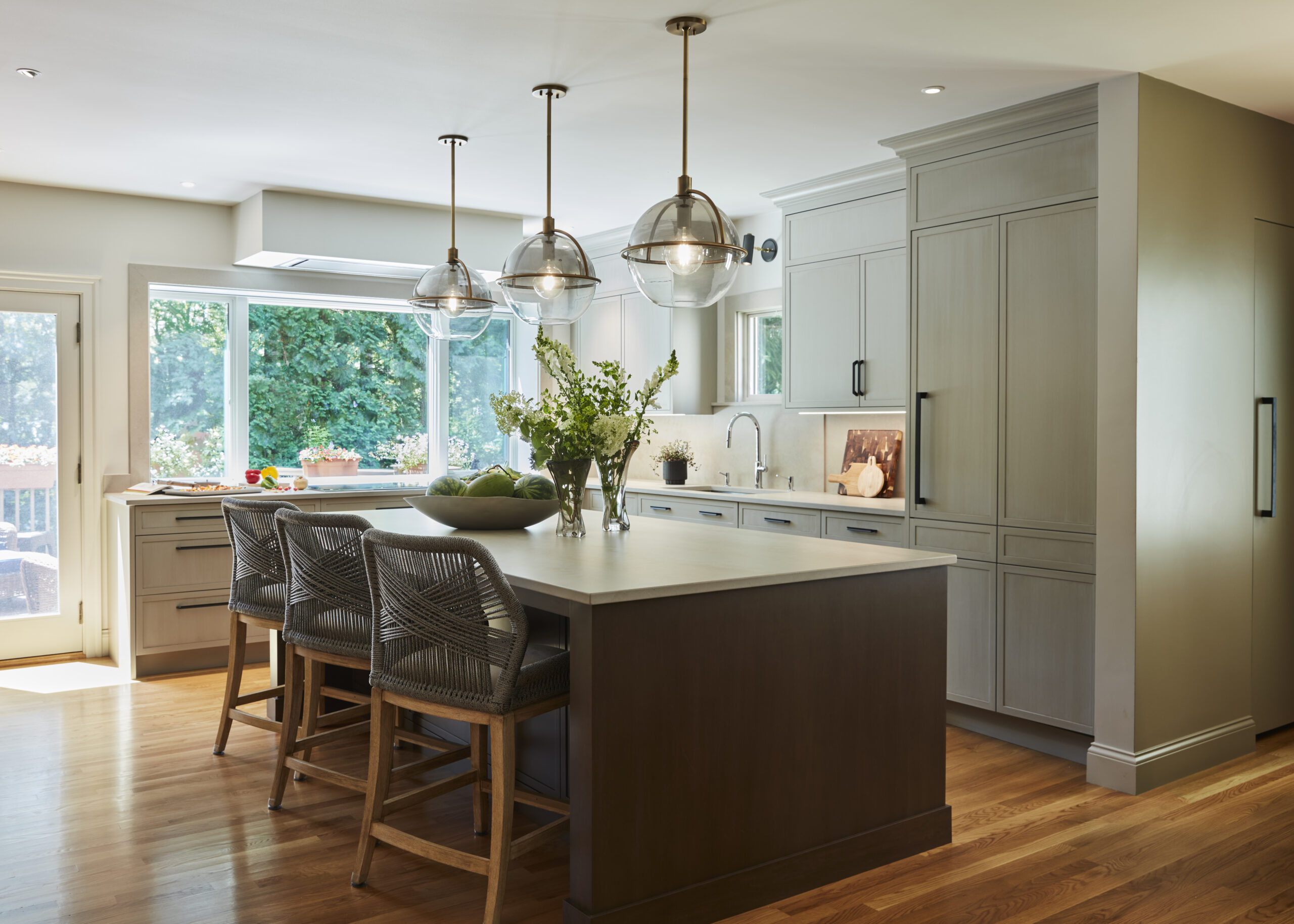 Modern kitchen with large island, three pendant lights, woven bar chairs, light gray cabinets, wood flooring, and a window overlooking greenery. Fresh flowers and fruit add a touch of Luxury Interior Design to the stylish space.