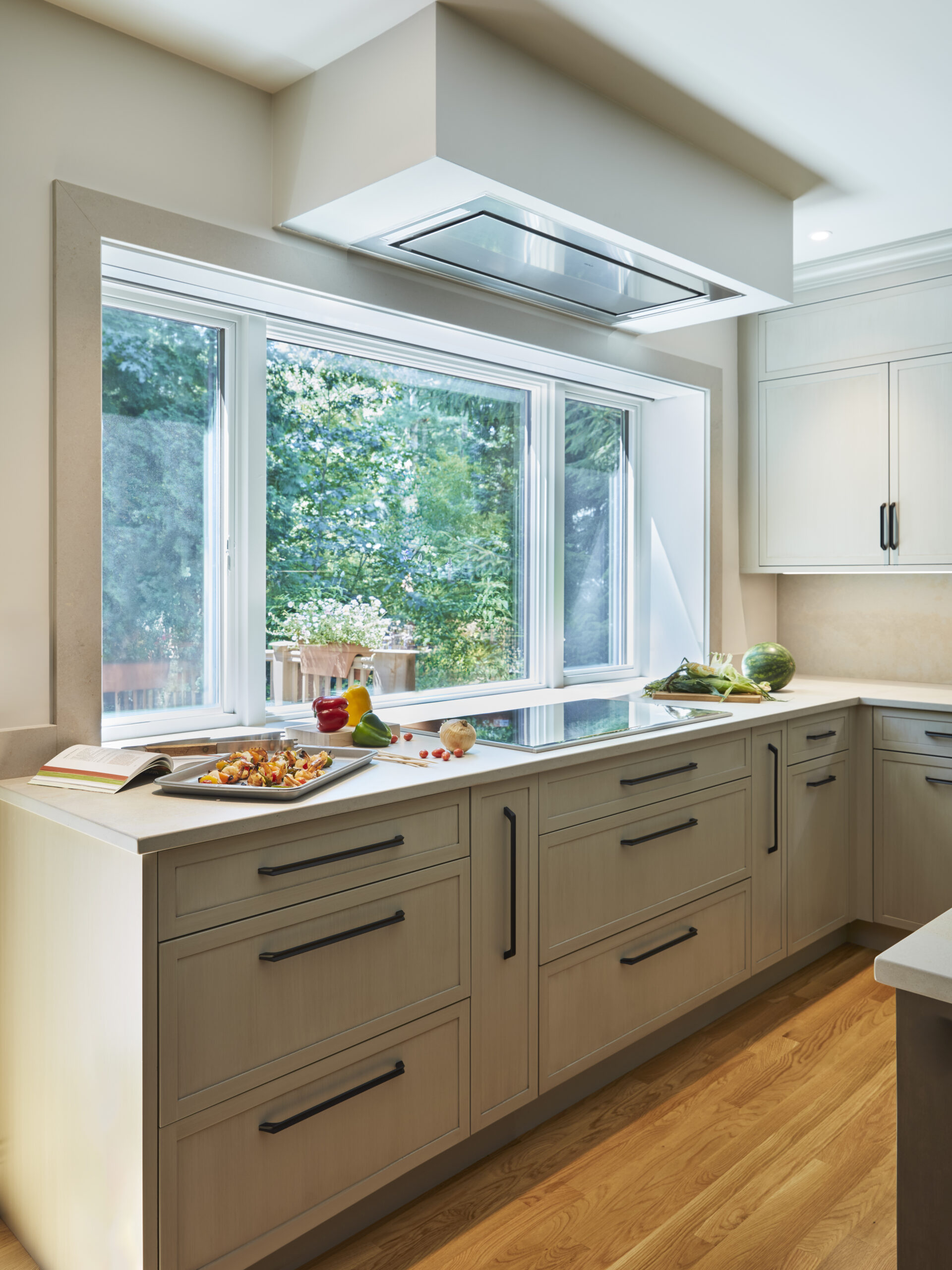 Modern kitchen with light wood cabinets, black handles, a large window overlooking greenery, and food items on the counter—showcasing luxury interior design by a top interior designer in Massachusetts.