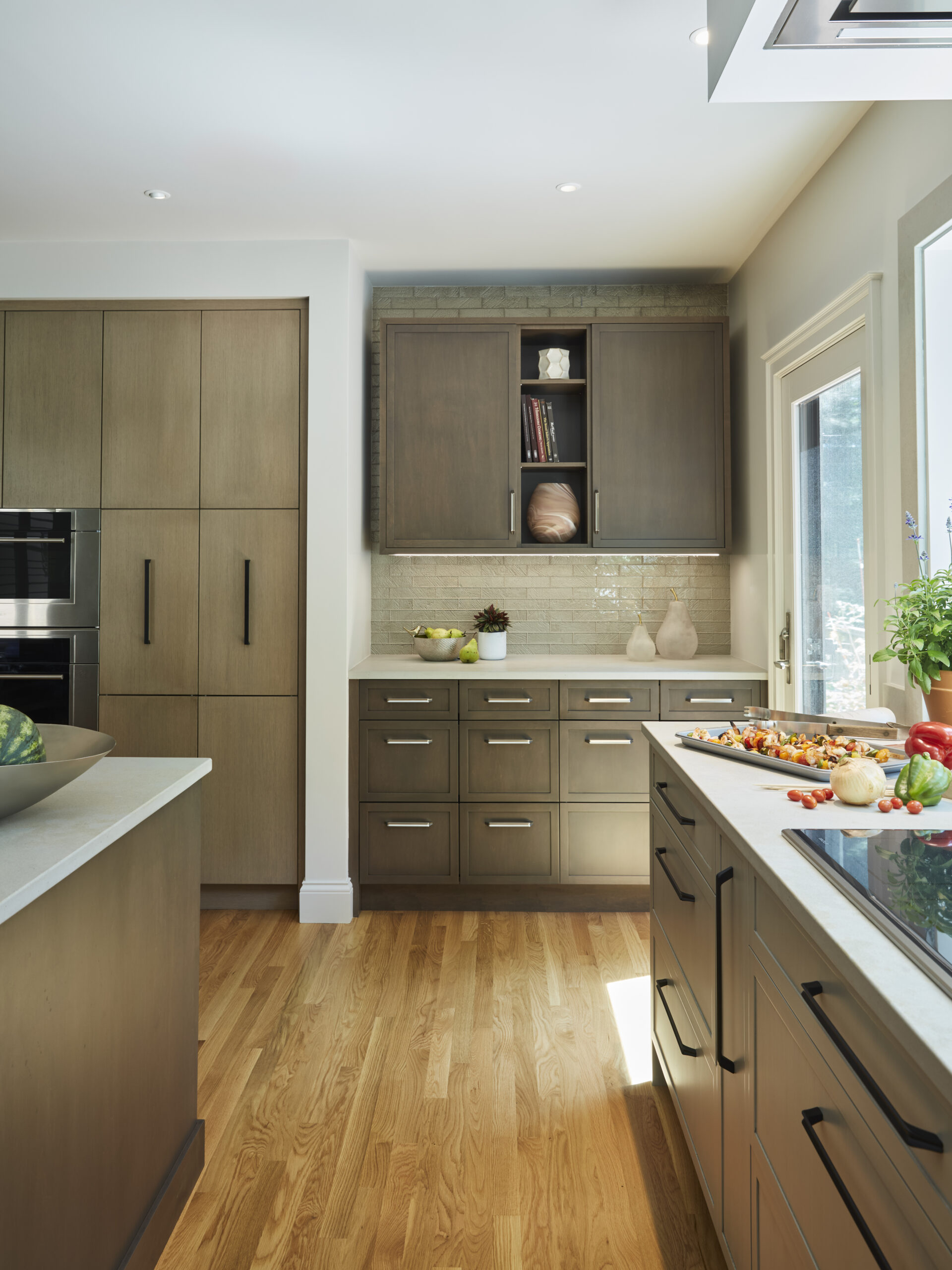 Modern kitchen with wooden cabinets, a light wood floor, and a large window; fresh vegetables and herbs are arranged on the countertop, showcasing elegant touches inspired by luxury interior design.