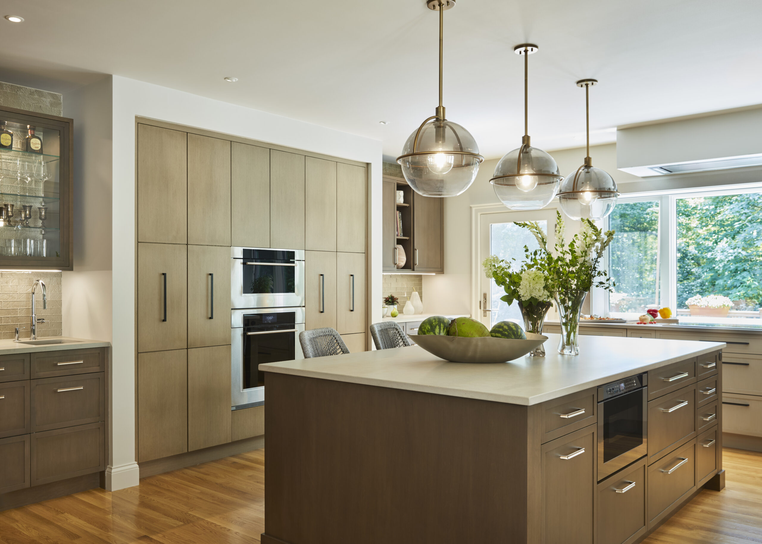 Modern kitchen with wooden cabinets, built-in ovens, a large island cooktop, pendant lights, barstools, and large windows filling the space with natural light—a perfect example of Luxury Interior Design by a top Interior Designer Massachusetts.