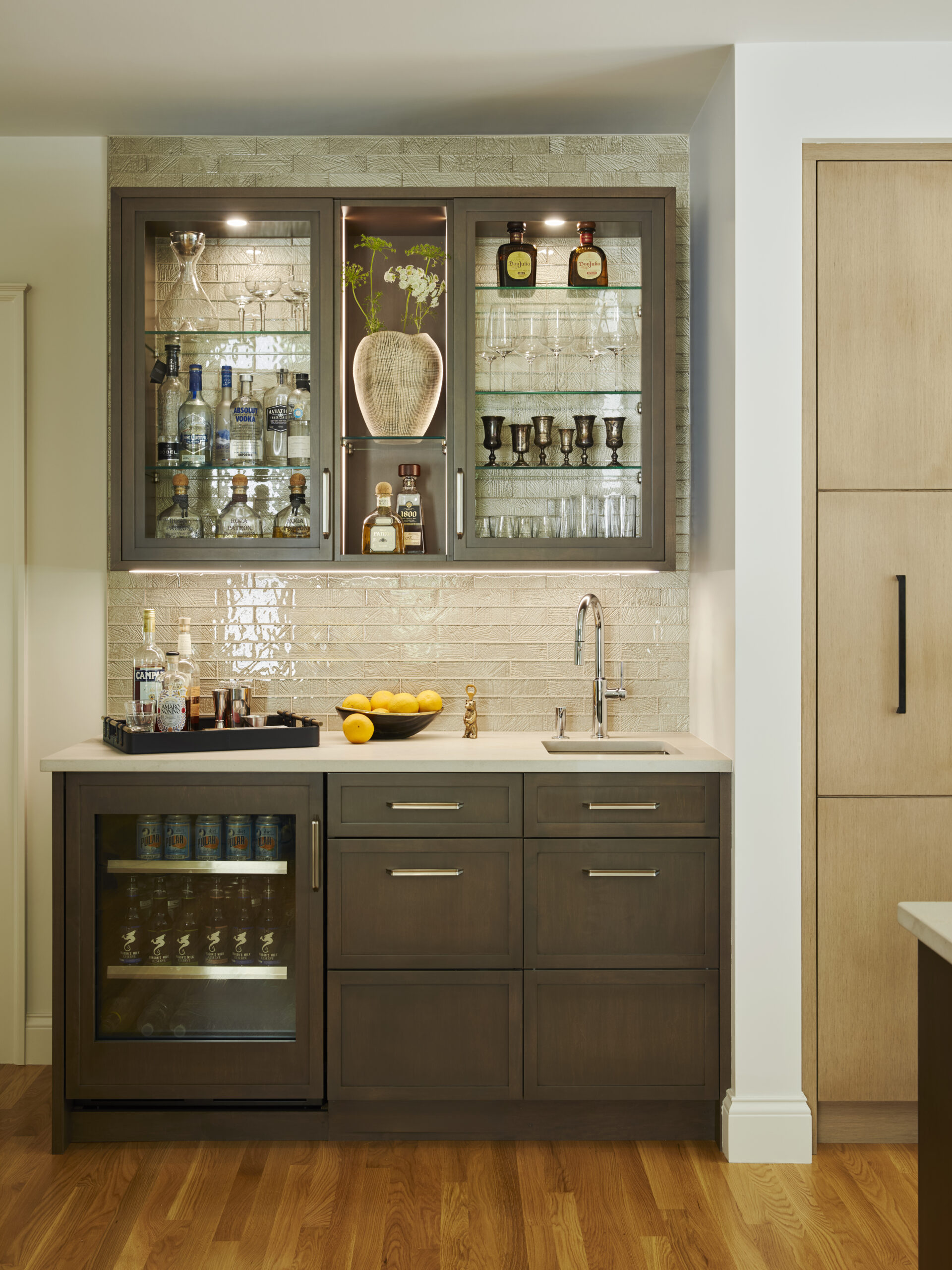 A small home bar setup designed with luxury interior design touches: glass cabinets display bottles and glassware, a countertop with lemons, a mini-fridge stocked with drinks, a sink, and elegant wooden drawers below.
