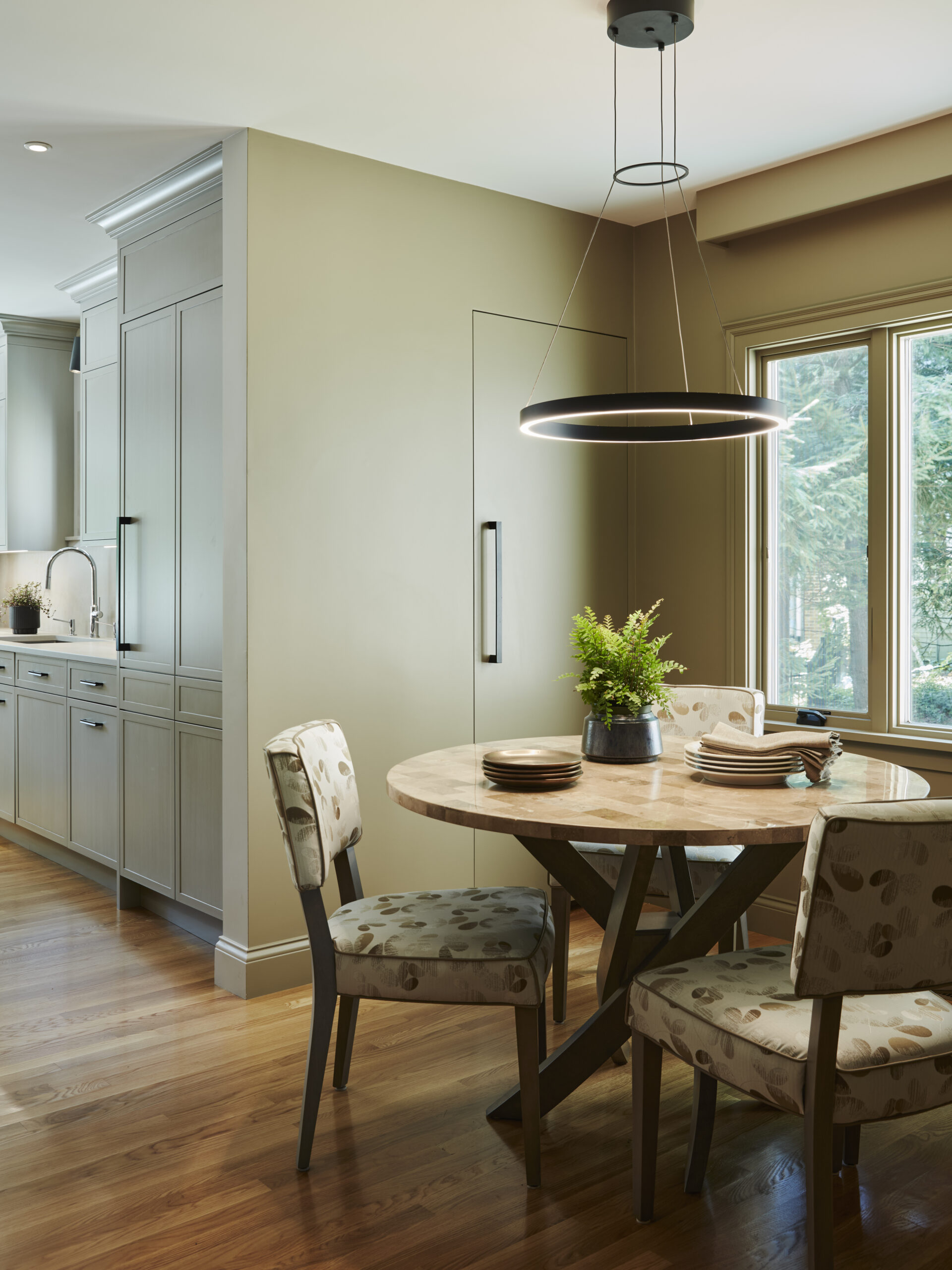 A round dining table with four patterned chairs, a potted plant centerpiece, and a modern circular light fixture create a luxury interior design vibe in this kitchen with wood floors and beige walls—perfect inspiration from an interior designer Massachusetts.