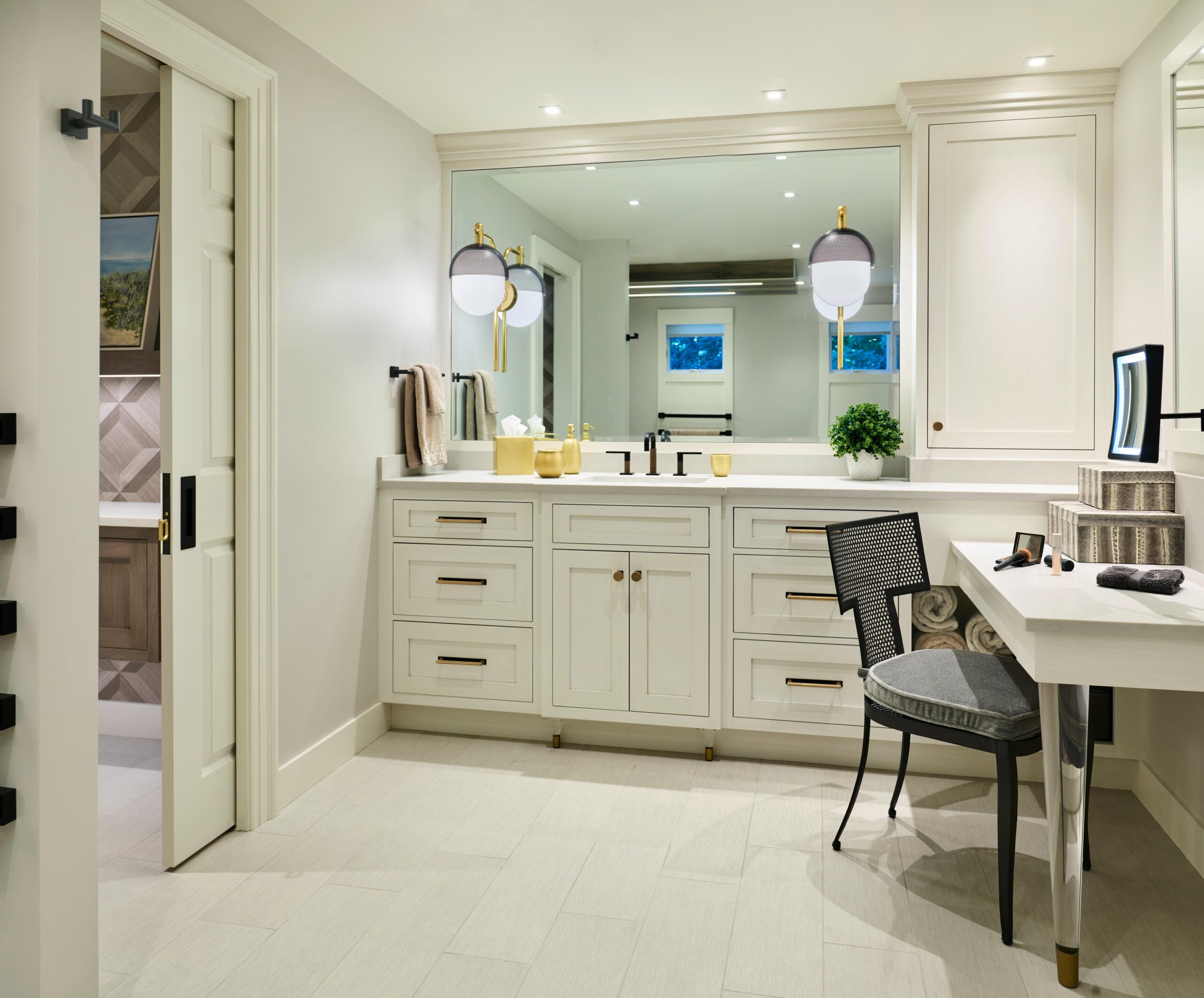 Modern bathroom with double sink vanity, large mirror, built-in cabinets, chair by a makeup counter, and neutral-colored decor with soft lighting.