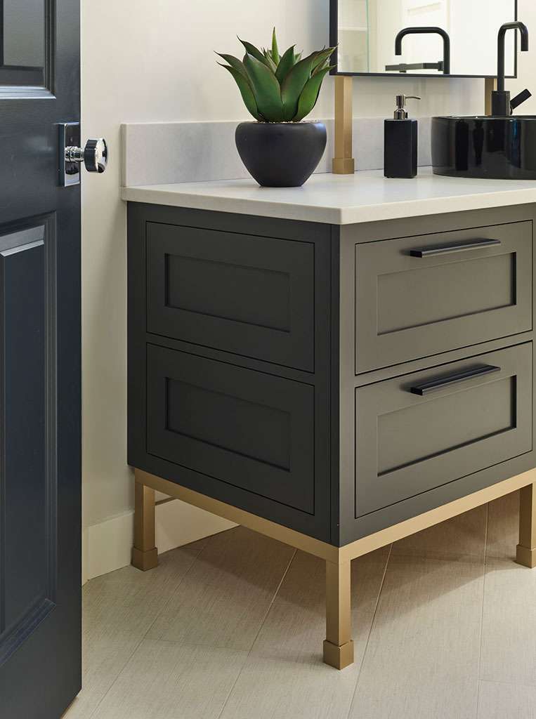 A modern bathroom vanity with dark drawers, gold legs, a white countertop, and a potted plant brings luxury interior design flair to any space, set against a light-colored wall and floor.