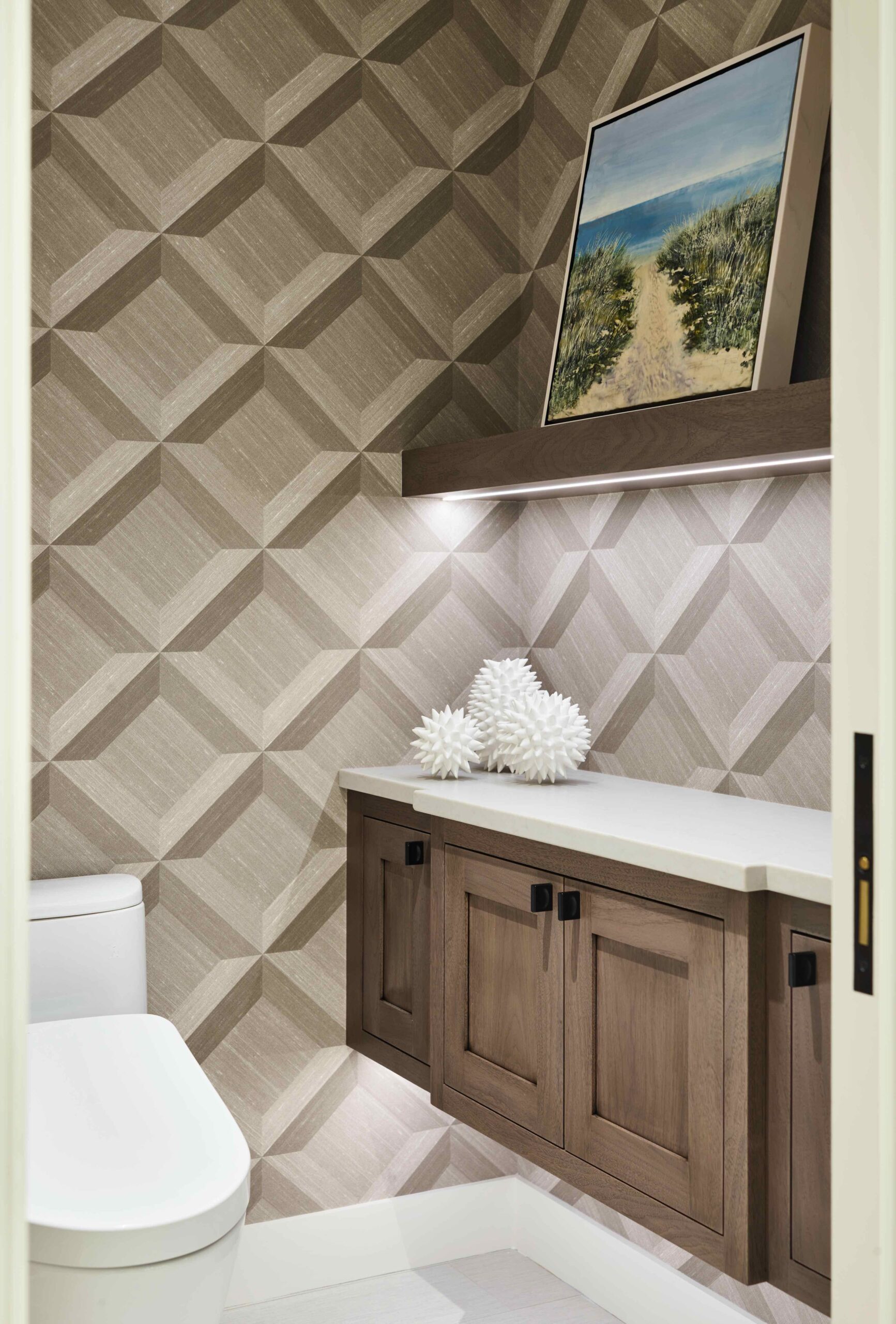 A modern bathroom with geometric-patterned wallpaper, a white toilet, wooden cabinets, decorative white coral objects, and a landscape painting on a shelf.