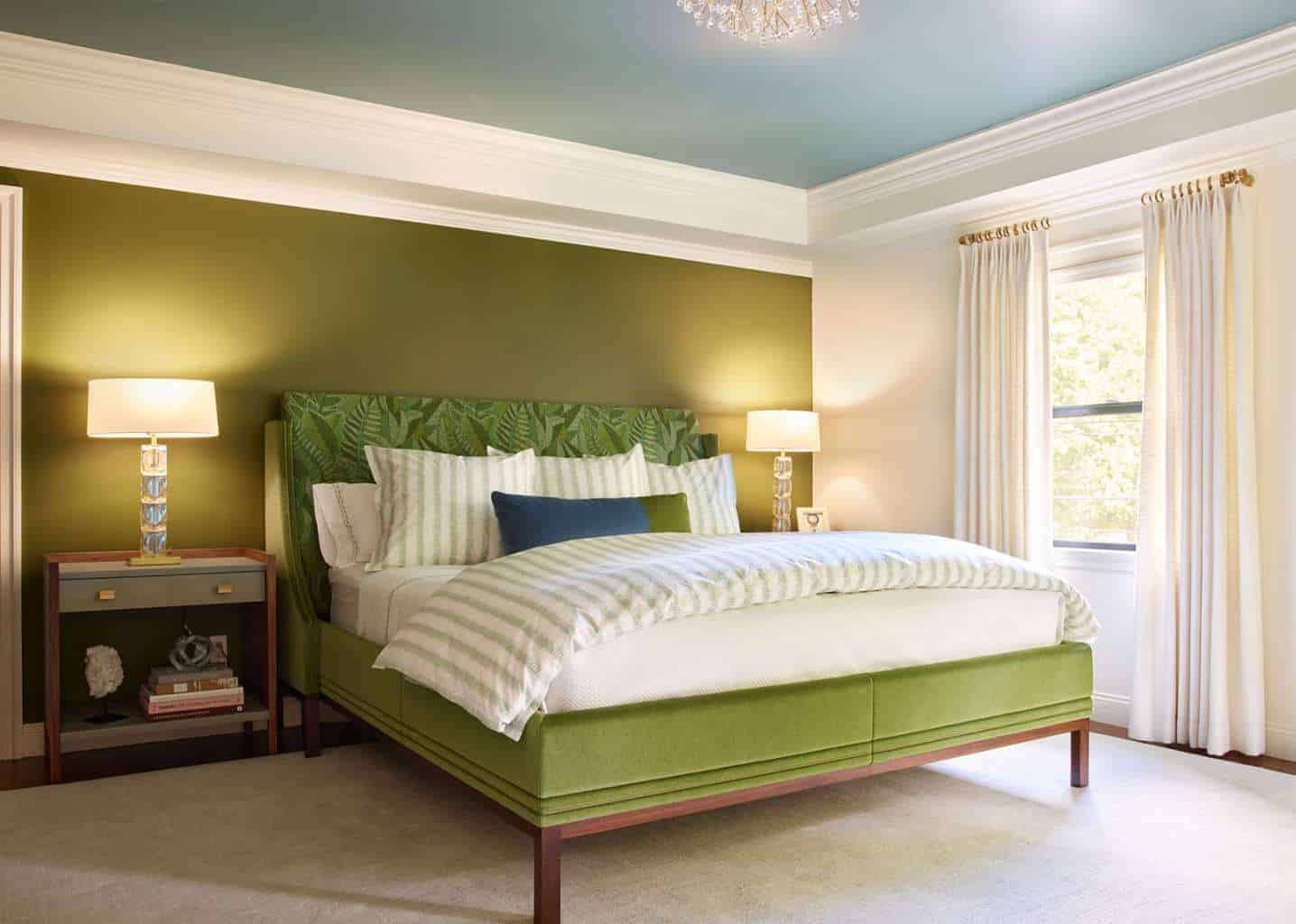 A bedroom by an expert Interior Designer Massachusetts features a green upholstered bed with striped bedding, nightstands with lamps, a green accent wall, large window with white curtains, and a blue ceiling—showcasing luxury interior design.