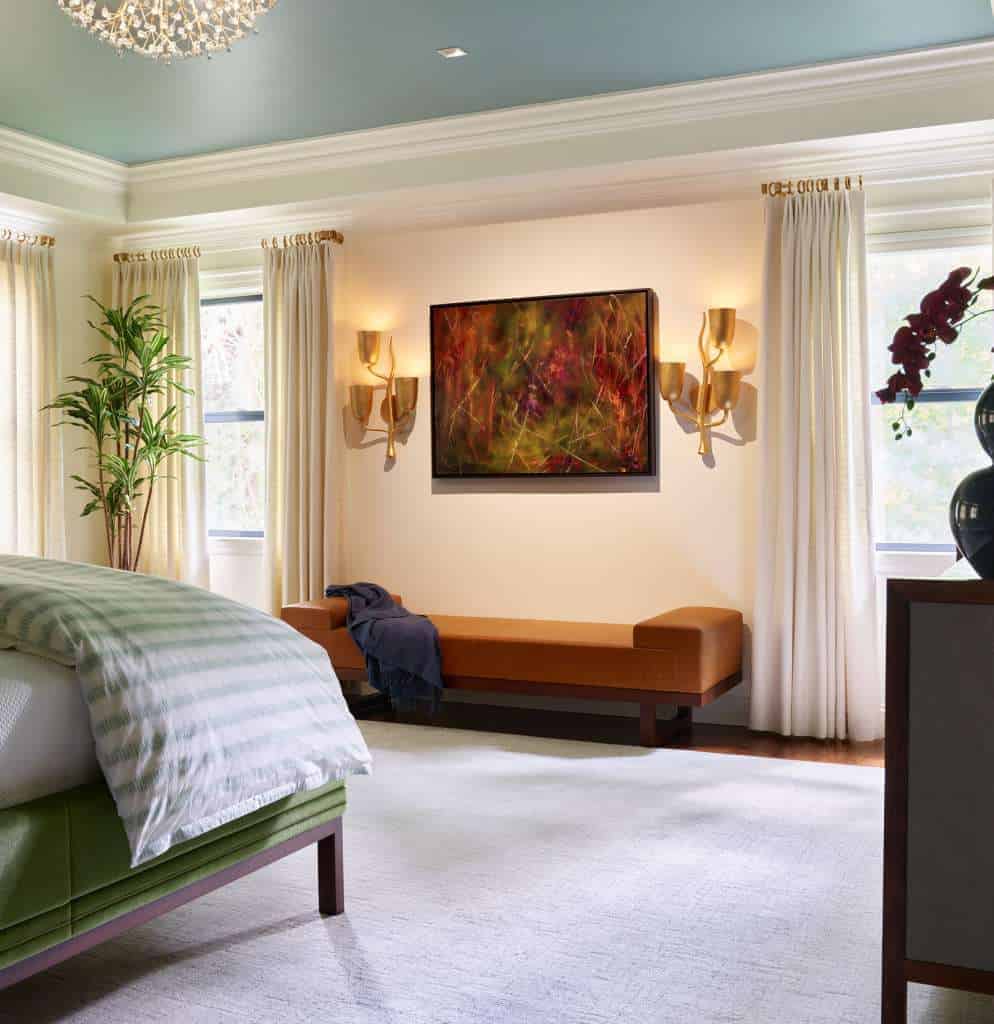 Bedroom with a green bed, orange bench, wall art, gold sconces, potted plant, and large windows with white curtains—an inviting space reflecting luxury interior design by a top interior designer in Massachusetts.