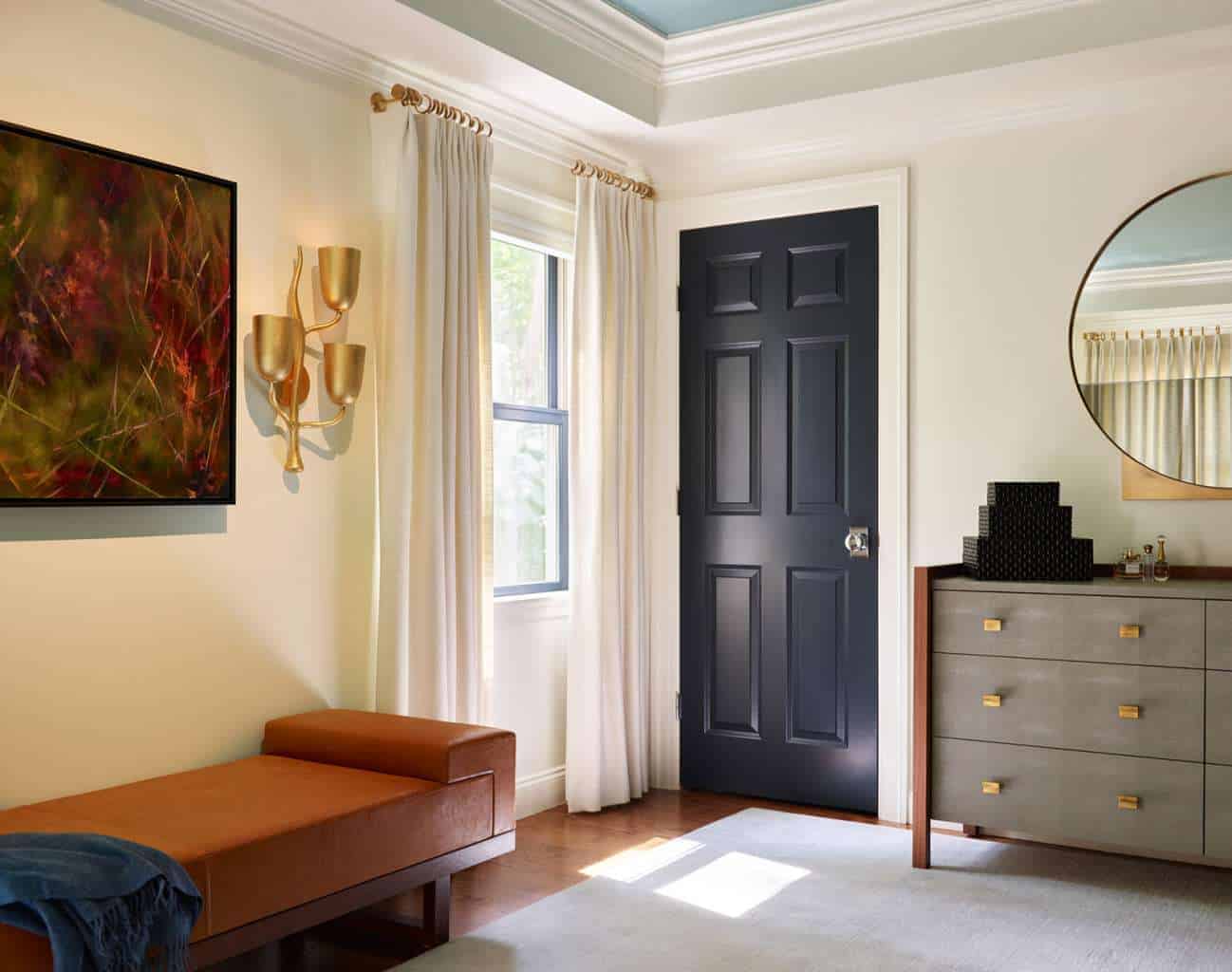 A modern room with luxury interior design featuring a black door, a round mirror above a gray dresser, a brown bench, white curtains, and abstract wall art.