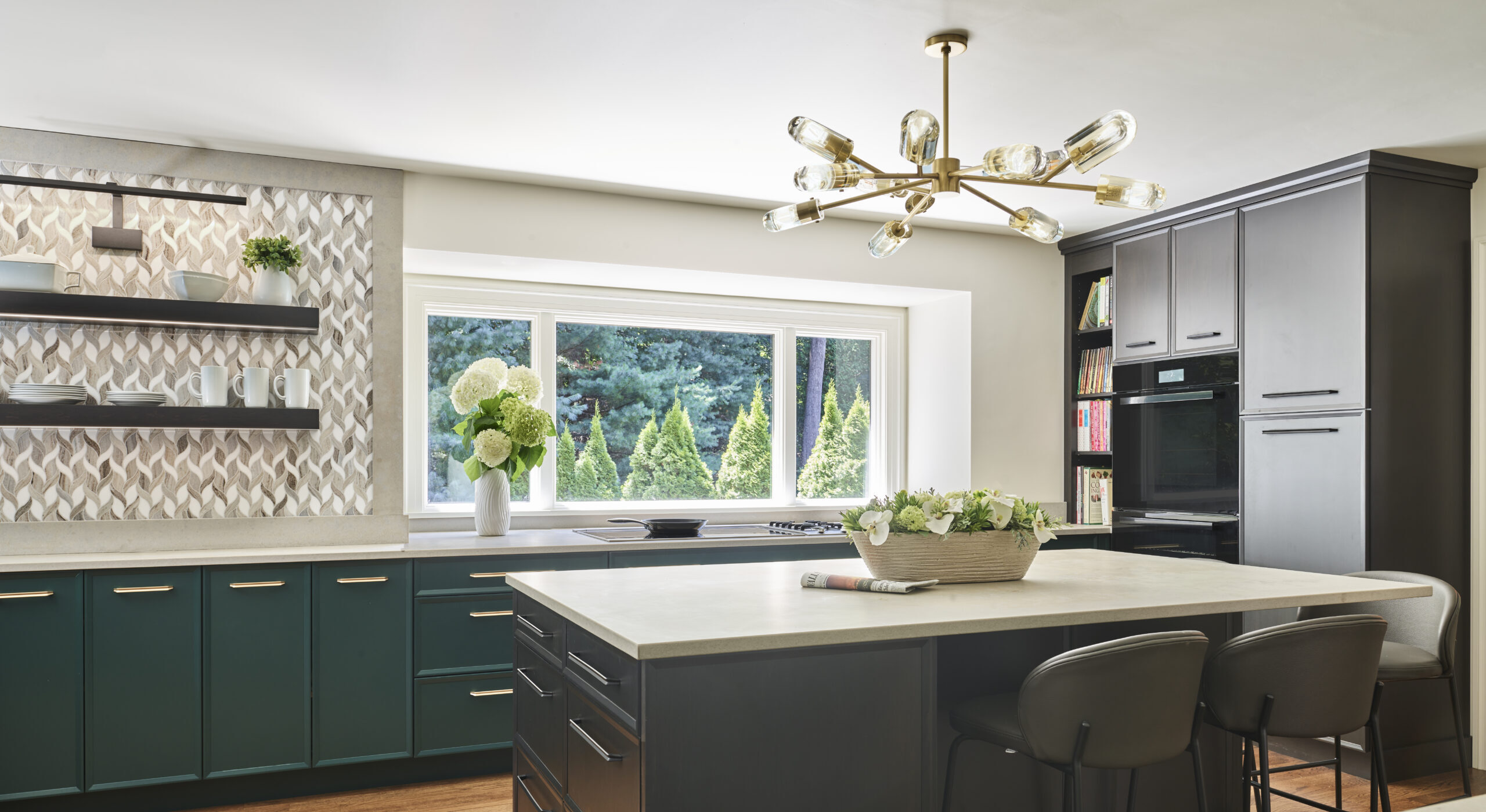 Modern kitchen with dark cabinets, large island with chairs, wall-mounted shelves, decorative backsplash, and a brass chandelier; window overlooks green trees—perfect for those seeking luxury interior design by an interior designer in Massachusetts.