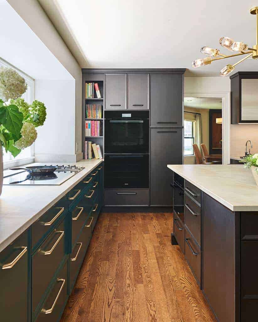 Modern kitchen with dark cabinets, white countertops, built-in oven, gas cooktop, bookshelves, hardwood floors, and a large window with flowers—showcasing the elegance of Luxury Interior Design by a top Interior Designer Massachusetts.
