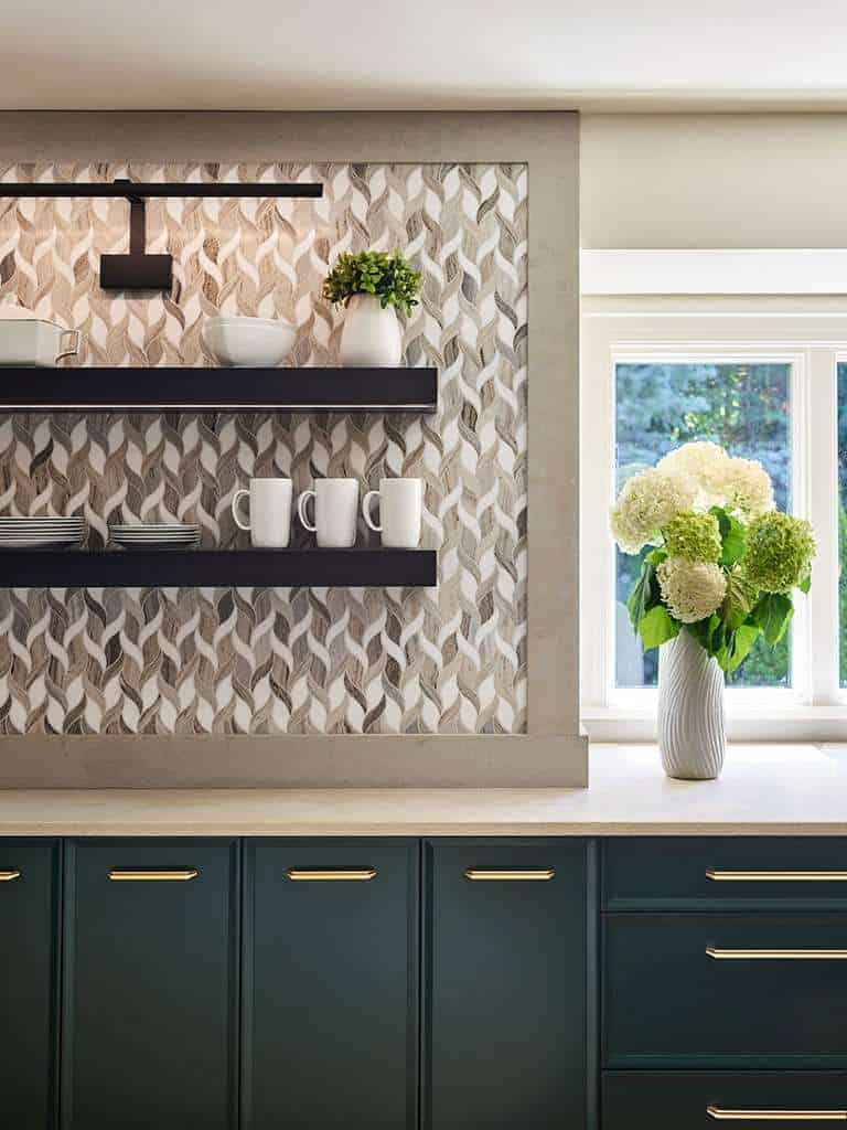 Modern kitchen with geometric tile backsplash, dark floating shelves holding white dishes and mugs, green cabinets with gold handles, and a vase of hydrangeas by a window—a perfect example of luxury interior design by an Interior Designer Massachusetts.