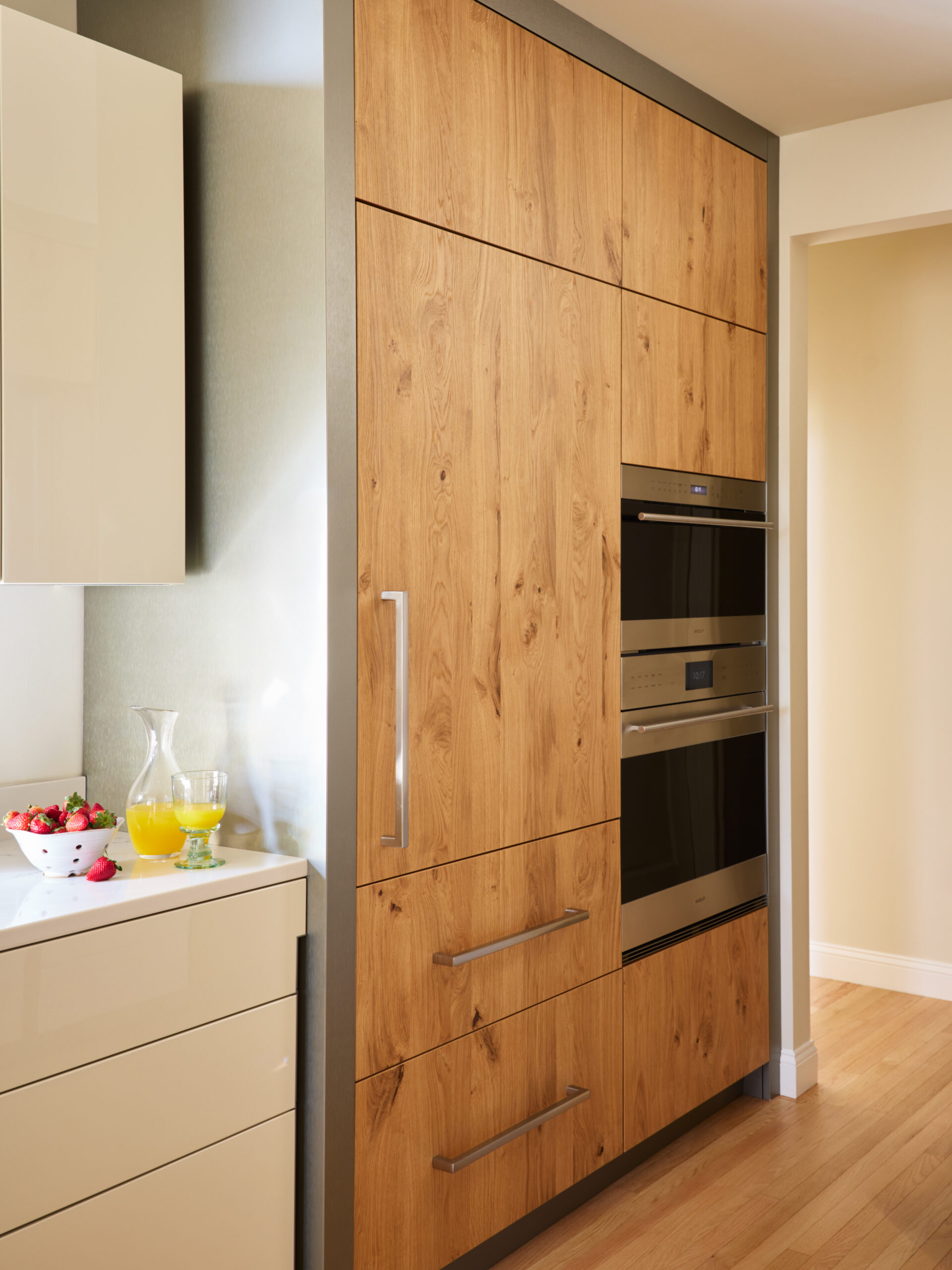 Modern kitchen with wood-paneled cabinets, built-in stainless steel oven and microwave, a white counter with a glass pitcher of orange juice, and a bowl of strawberries—perfect for luxury interior design in Massachusetts.
