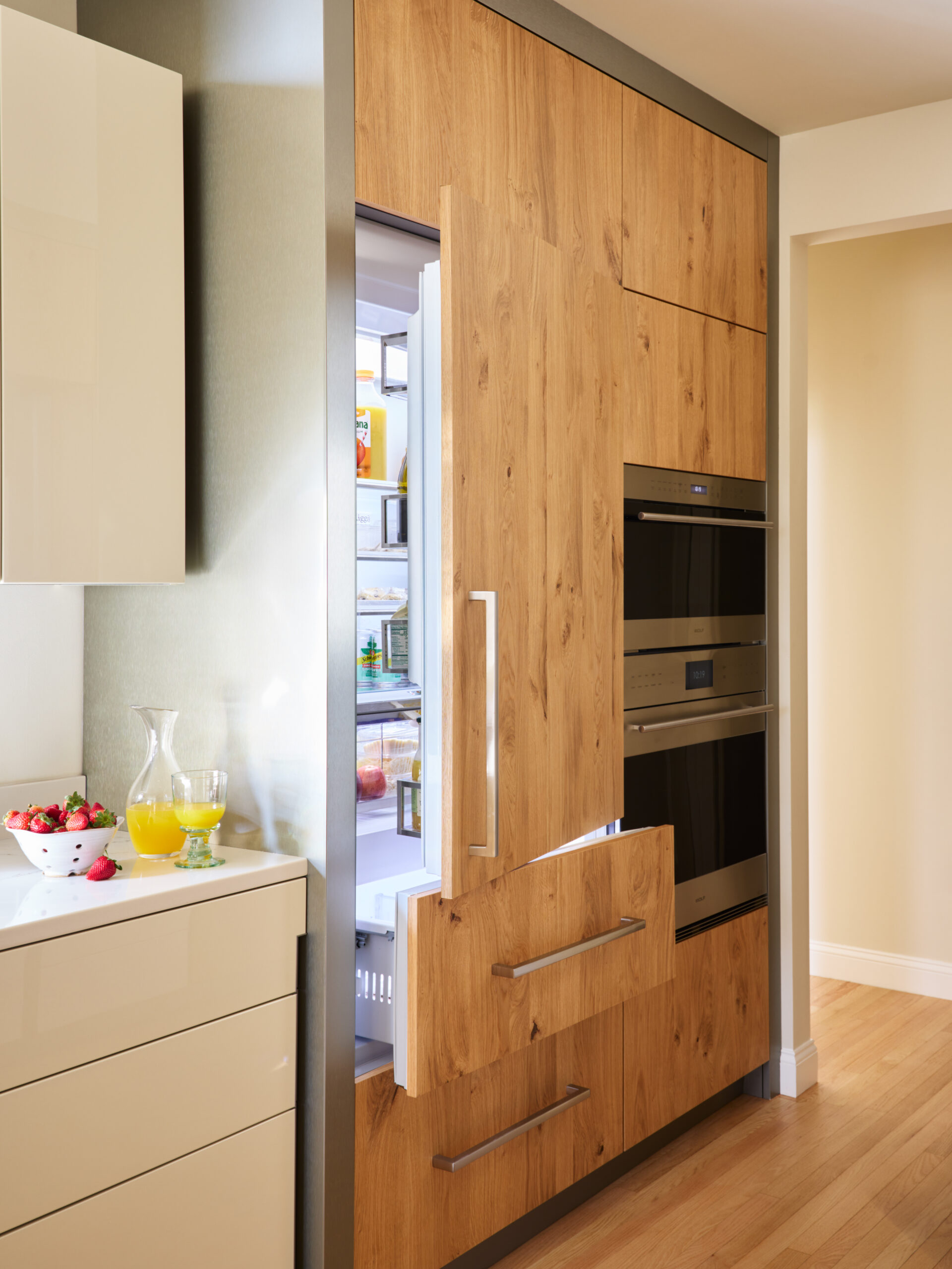 A modern kitchen with built-in wooden cabinets, an open refrigerator and freezer, integrated oven, and a countertop with juice, a glass, and a bowl of strawberries—perfectly showcasing luxury interior design.