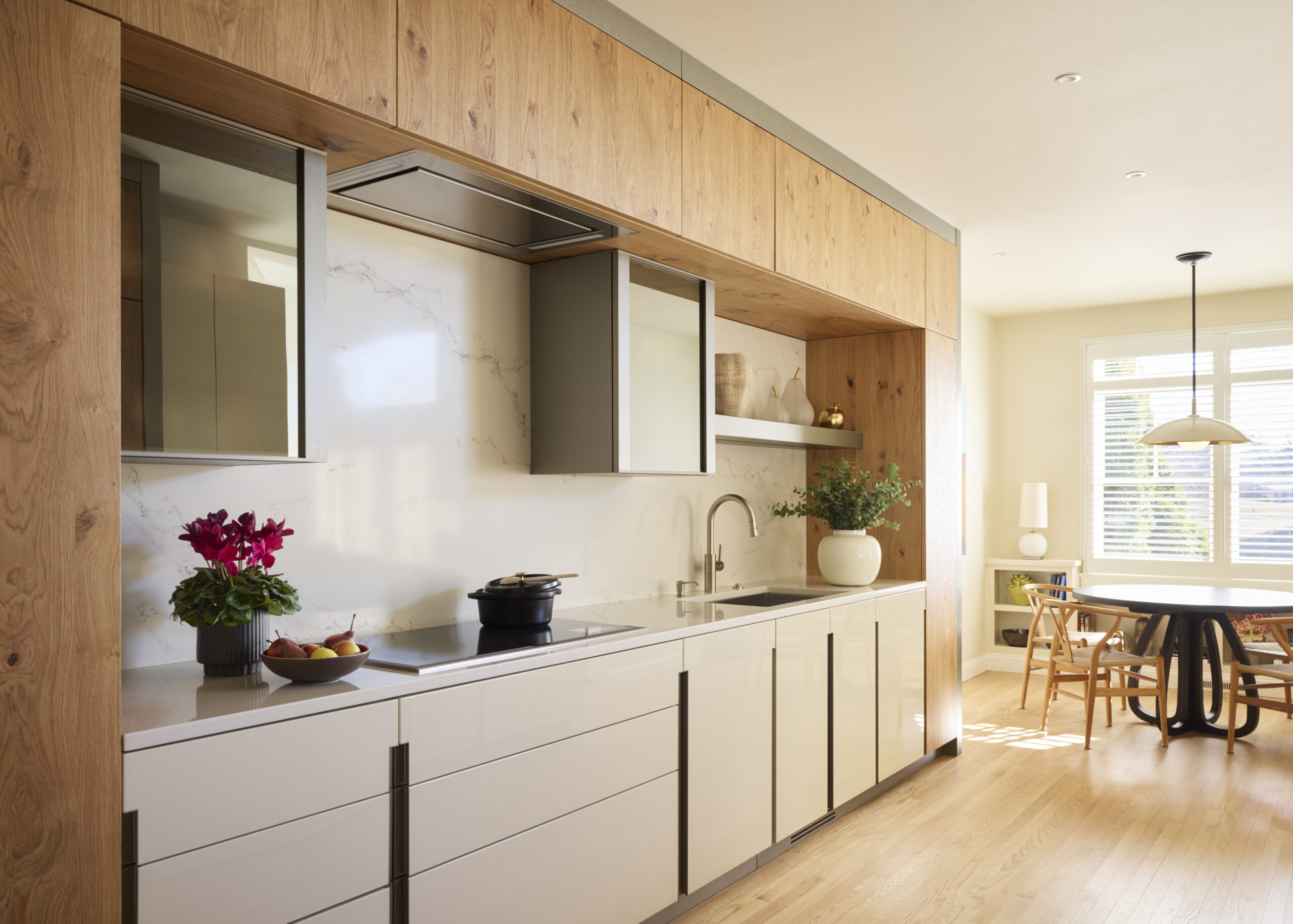 Modern kitchen with light wood cabinets, white countertops, and built-in appliances, styled by an Interior Designer Massachusetts. Dining area features a round table by the window and potted plants for a touch of luxury interior design.