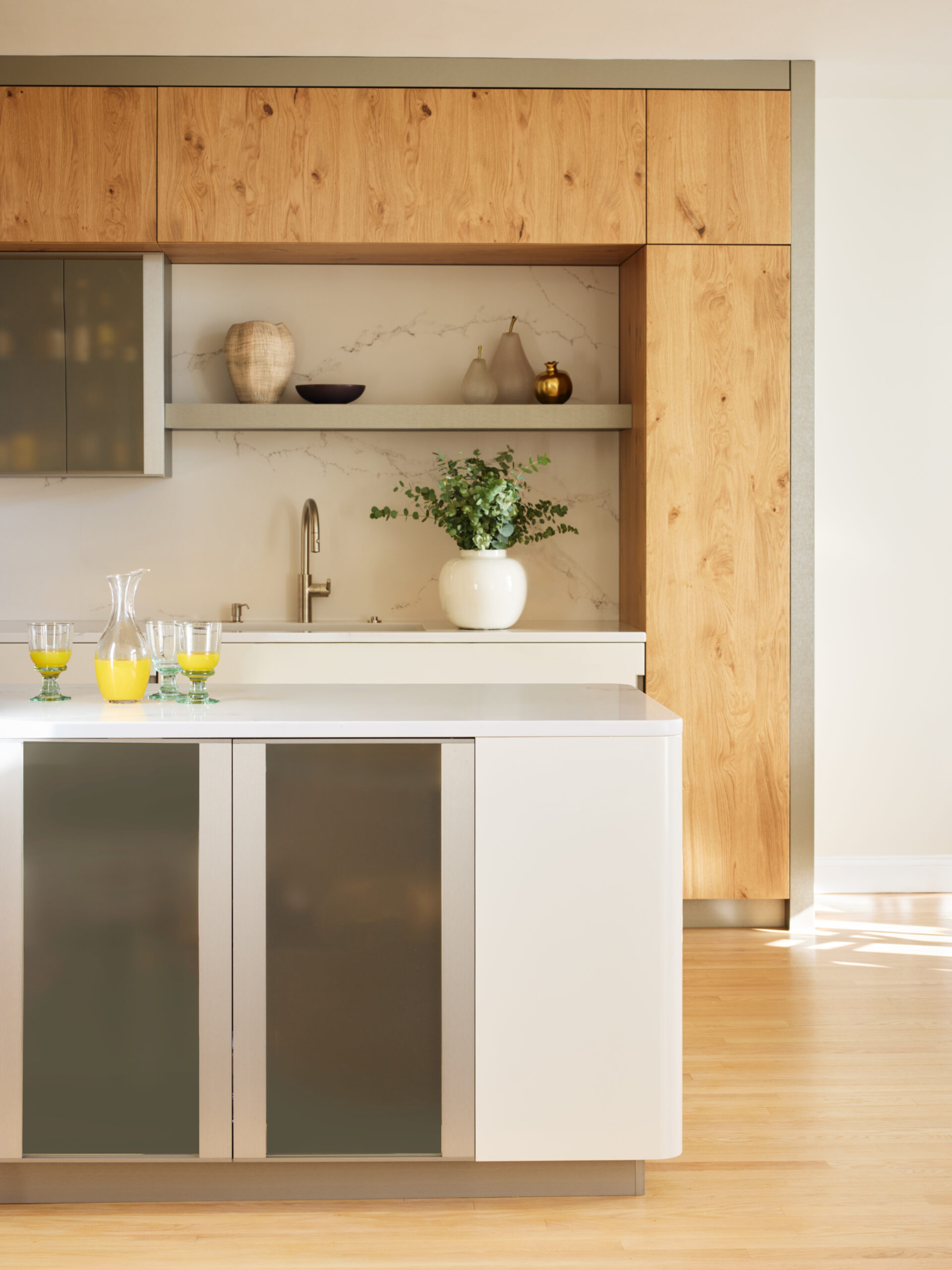 Modern kitchen with wood cabinets, a white countertop island, glassware filled with orange juice, potted plant, and decorative items on shelves against a marble backsplash—an example of luxury interior design by top interior designers in Massachusetts.