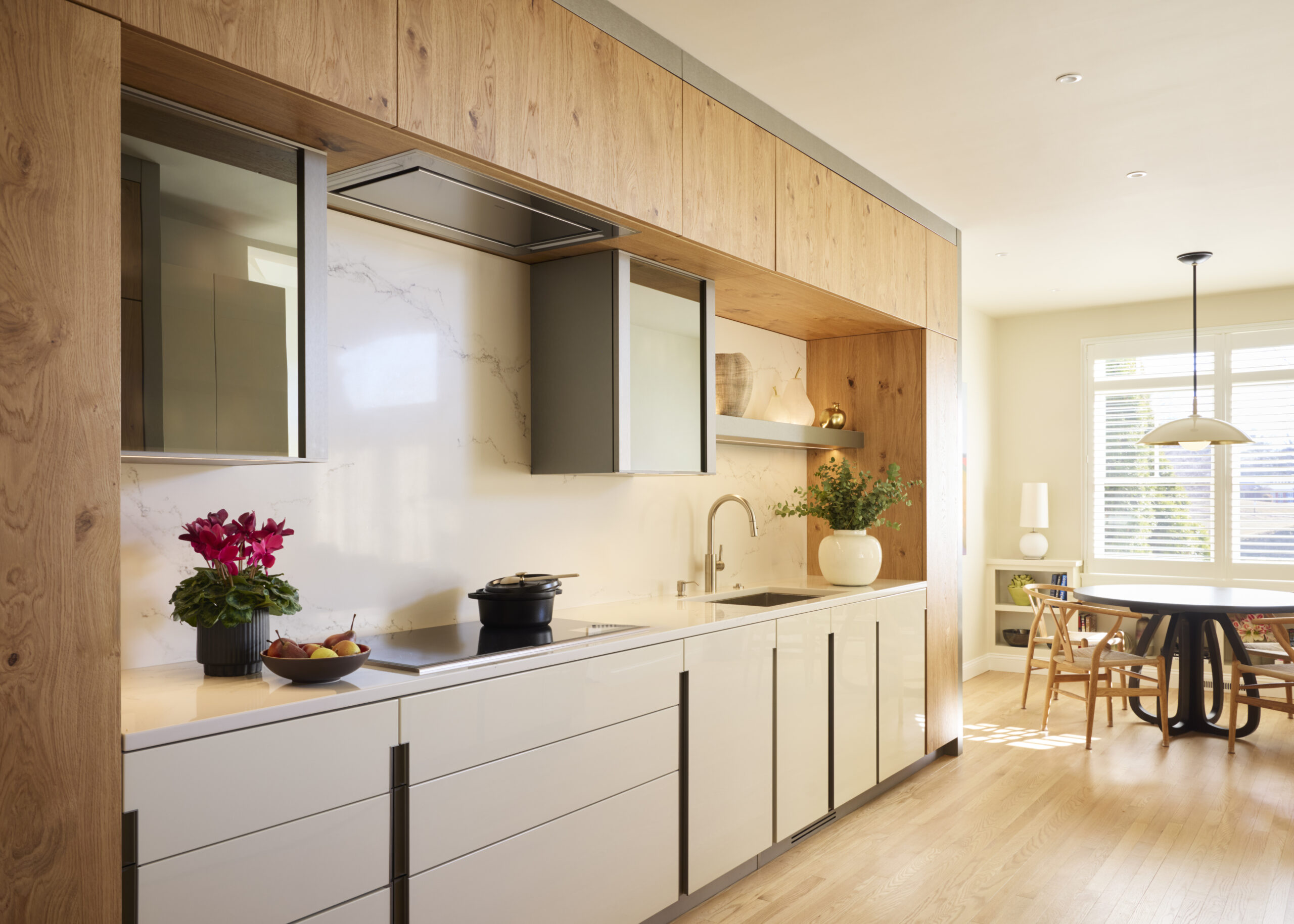 Modern kitchen with wood and white cabinetry, marble backsplash, built-in appliances, and decorative plants; luxury interior design details enhance the space, while the dining area with round table and chairs is visible in the background.