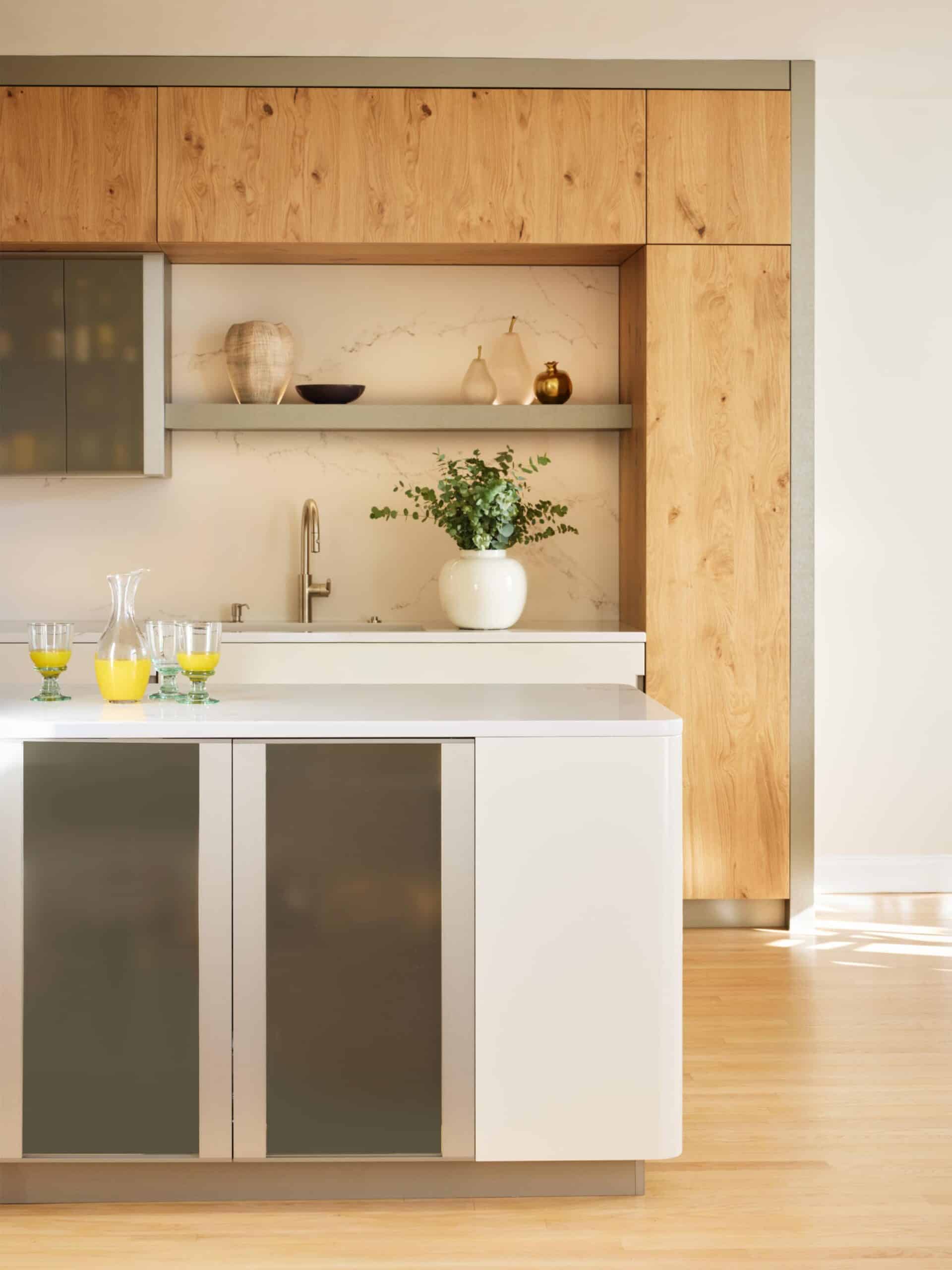 Modern kitchen with wood cabinets, a white island, glassware with orange juice, a sink, a vase with greenery, and decorative items on a shelf against a marble backsplash.