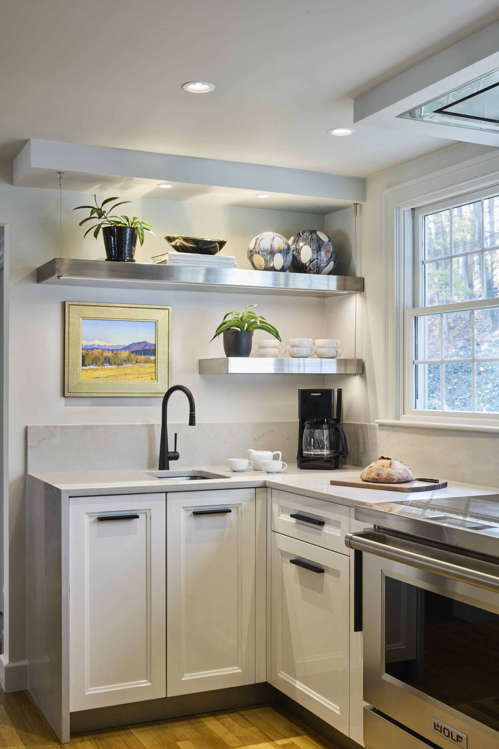Modern kitchen corner with white cabinets, black faucet, open shelves holding decor and plants—a perfect example of Luxury Interior Design by an Interior Designer Massachusetts. Natural light pours in through the window, highlighting a bread loaf and coffee maker.