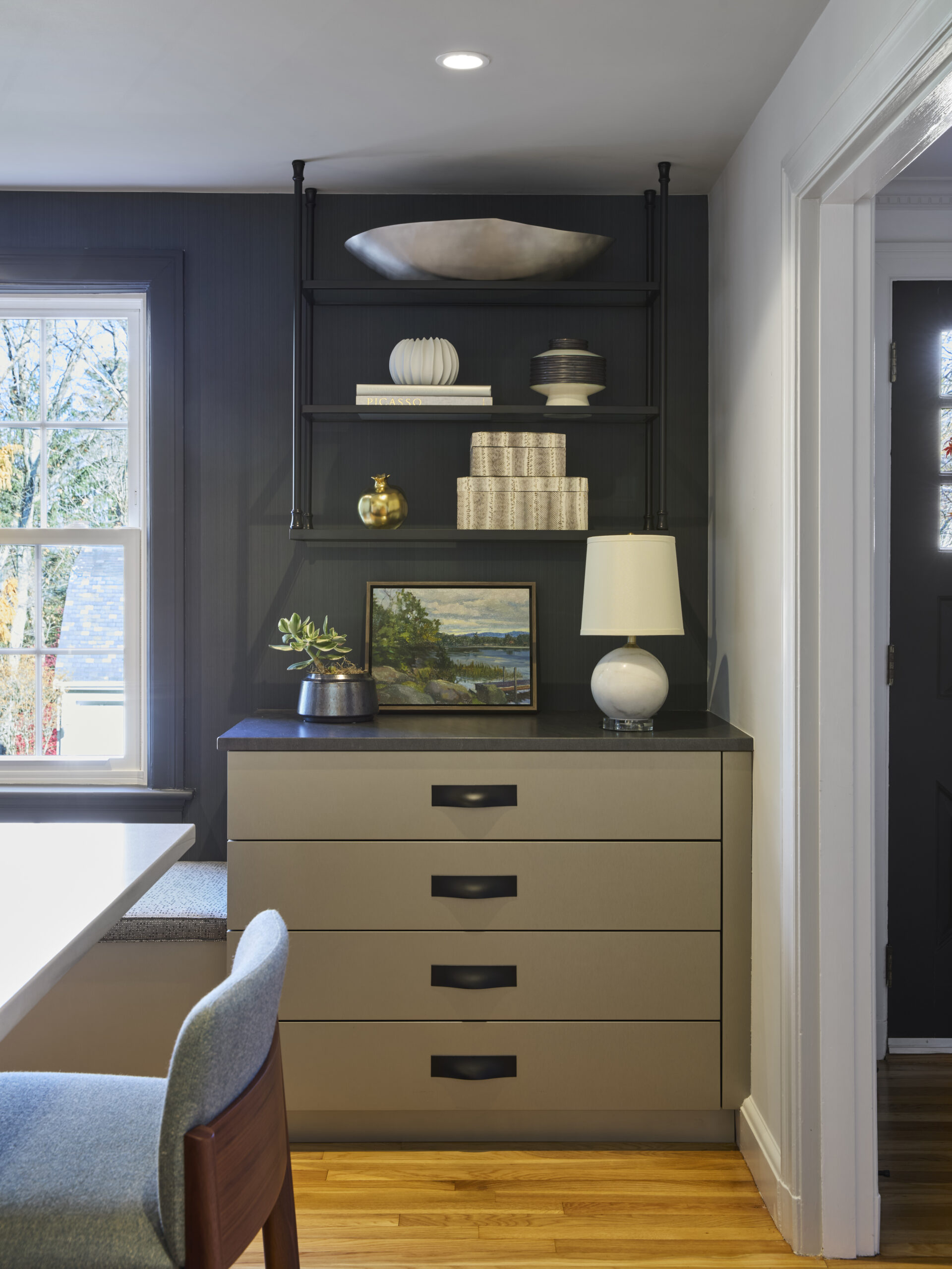 A modern corner with gray drawers, black shelves holding decor items, a lamp, framed artwork, and sunlight streaming in—perfect inspiration for luxury interior design or any interior designer in Massachusetts.