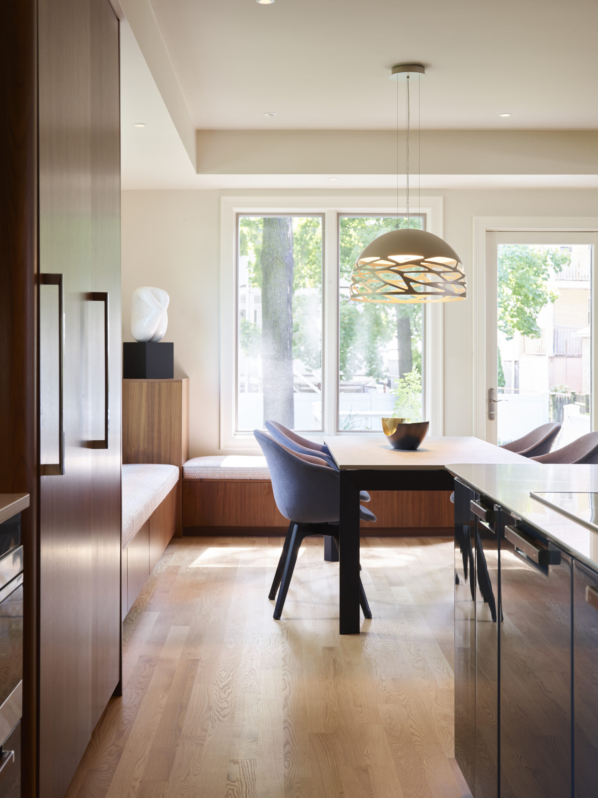 Experience luxury interior design in this modern kitchen and dining area with wooden floors, a sleek black table, blue chairs, built-in bench seating, pendant light, and large windows flooding the space with natural light.