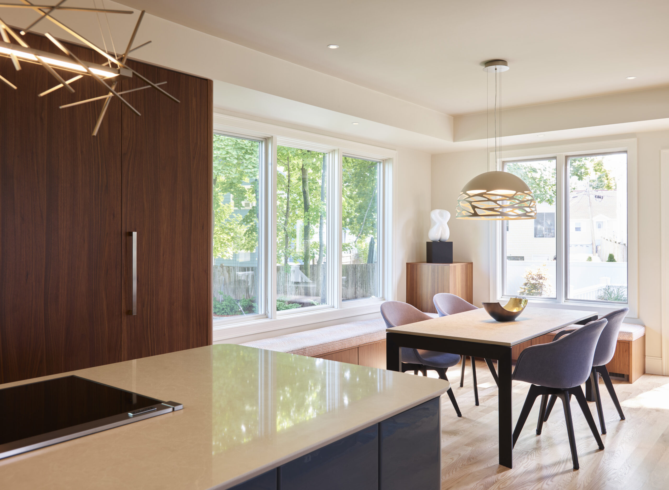 Modern dining area with a wooden table, four gray chairs, large windows, and contemporary pendant lights in a bright, minimalist interior by a top Interior Designer Massachusetts specializing in luxury interior design.