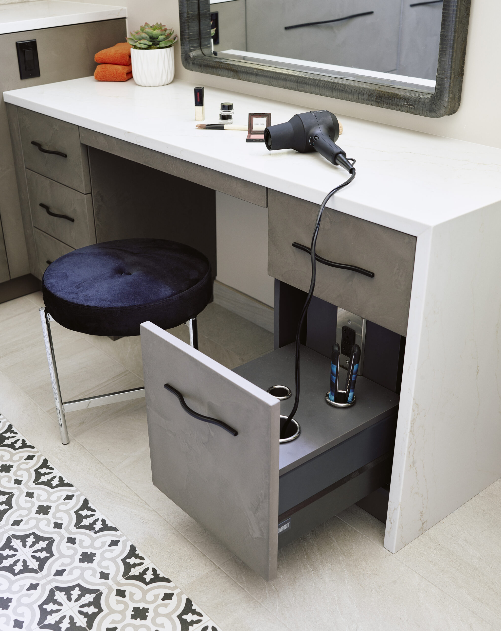 A modern vanity with a marble countertop, open drawer holding a hair dryer and styling tools, cushioned stool, and patterned floor tiles—perfect for those who appreciate luxury interior design crafted by an expert interior designer in Massachusetts.