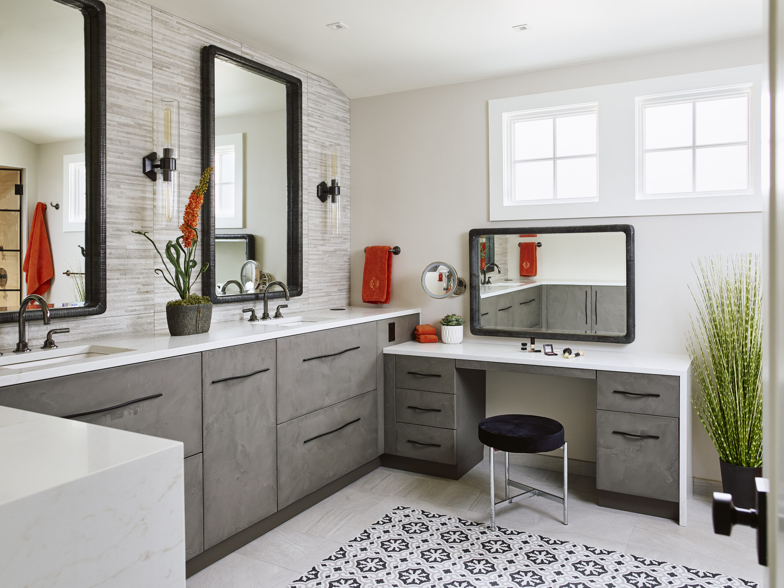 Modern bathroom with double sinks, a makeup vanity with mirror and stool, gray cabinets, decorative plants, and black and white patterned floor tile—expertly curated by a leading Luxury Interior Design firm in Massachusetts.