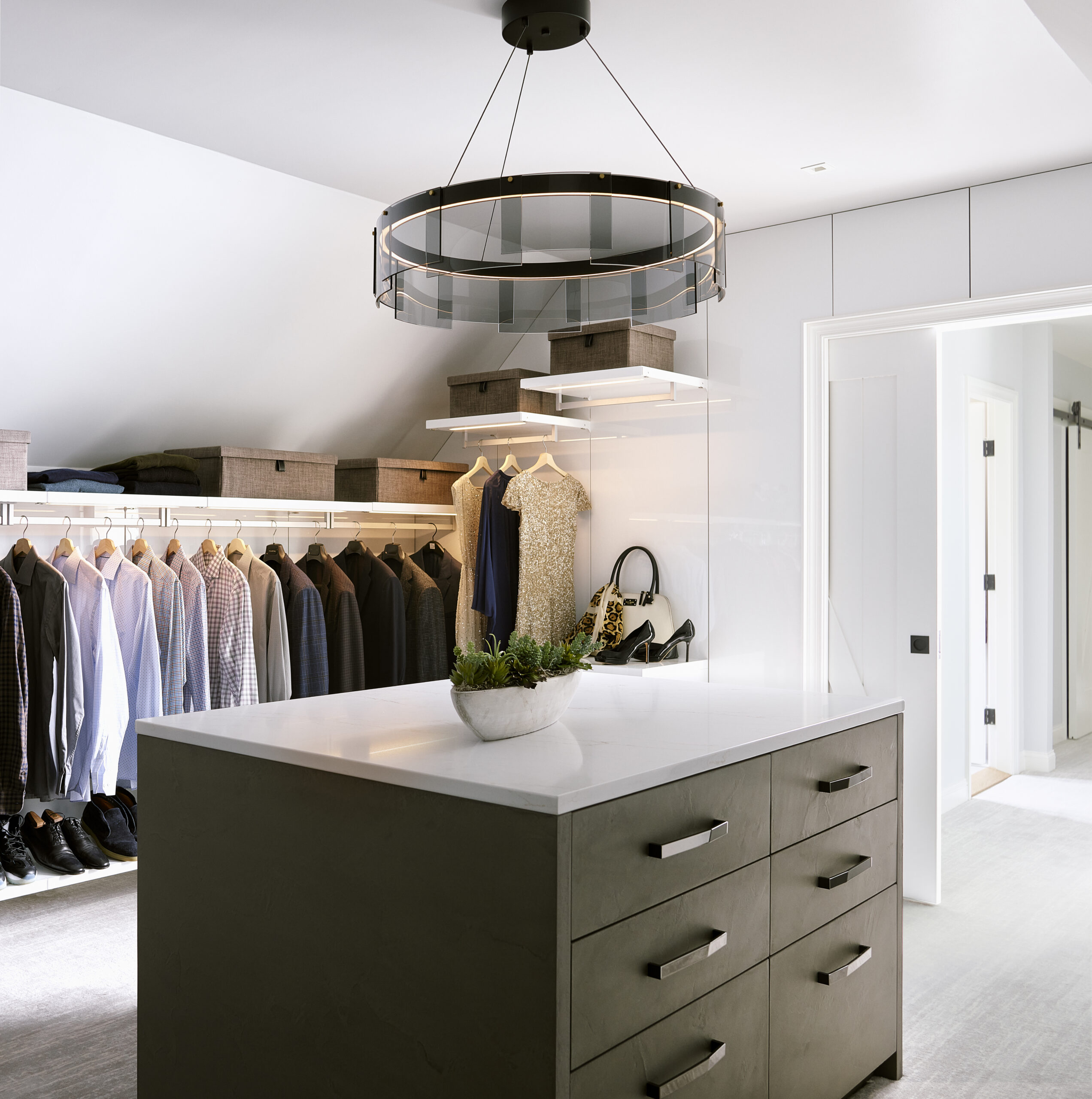 Modern walk-in closet with a central island, hanging clothes, storage boxes, handbags, and a large round ceiling light fixture—perfect for those seeking luxury interior design crafted by an expert interior designer in Massachusetts.