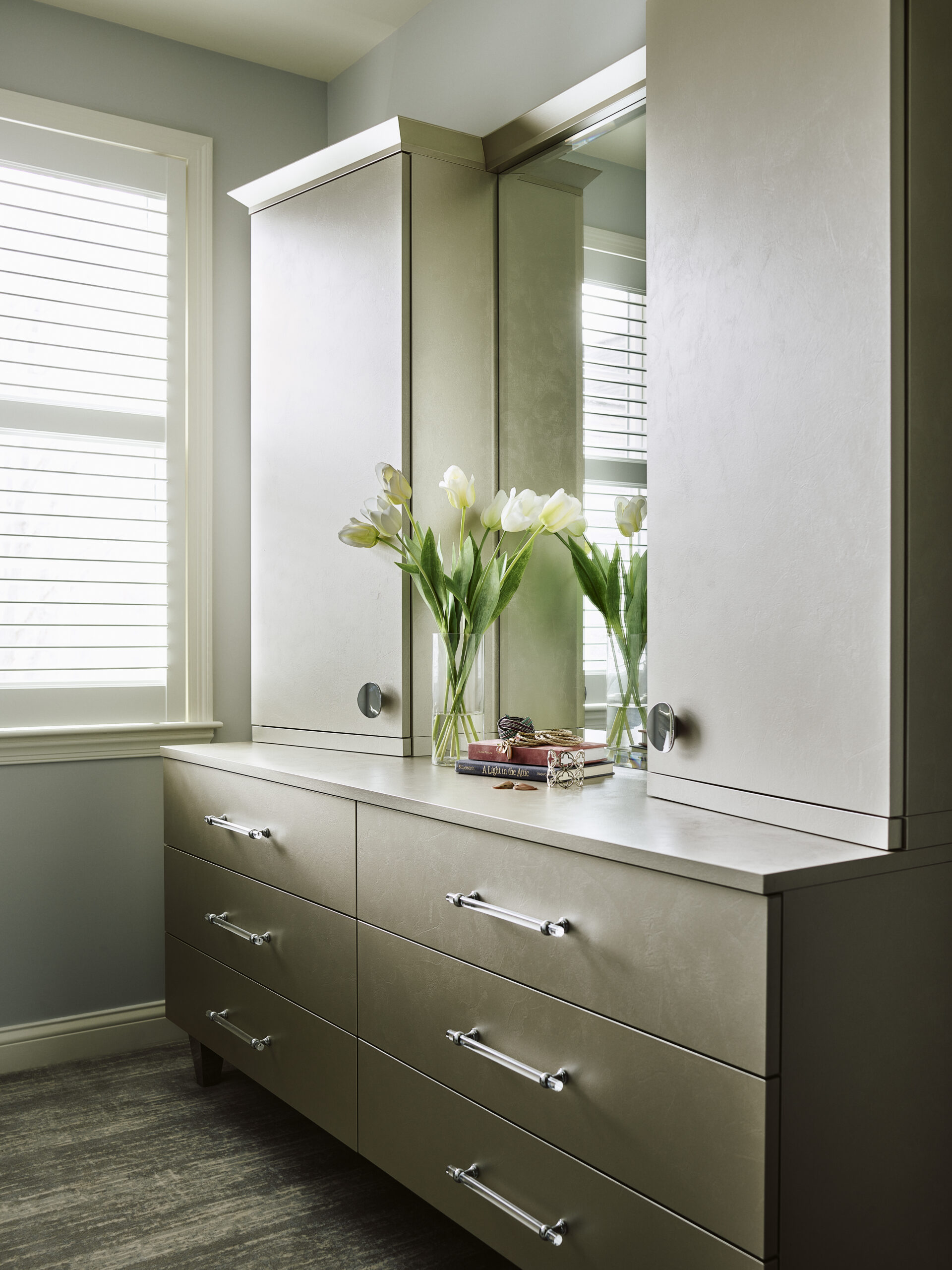 A beige dresser with silver handles stands below a large mirror, styled by a Luxury Interior Design expert. White tulips, books, and decor grace the top, while a window with blinds to the left completes this elegant Massachusetts space.
