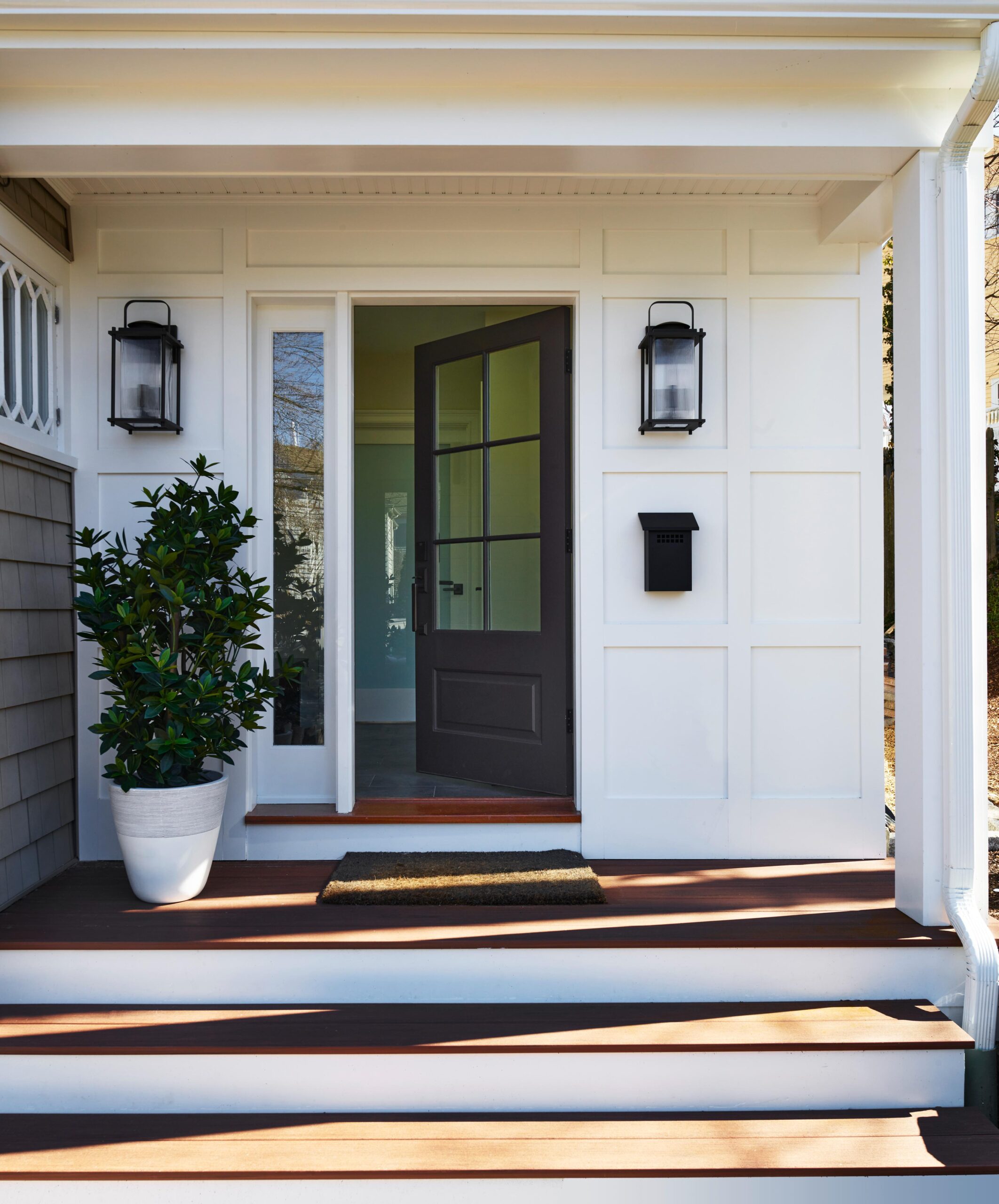 White front porch with steps, a black door partly open, a potted plant on the left, two black wall lanterns, and a black mailbox mounted on the wall.