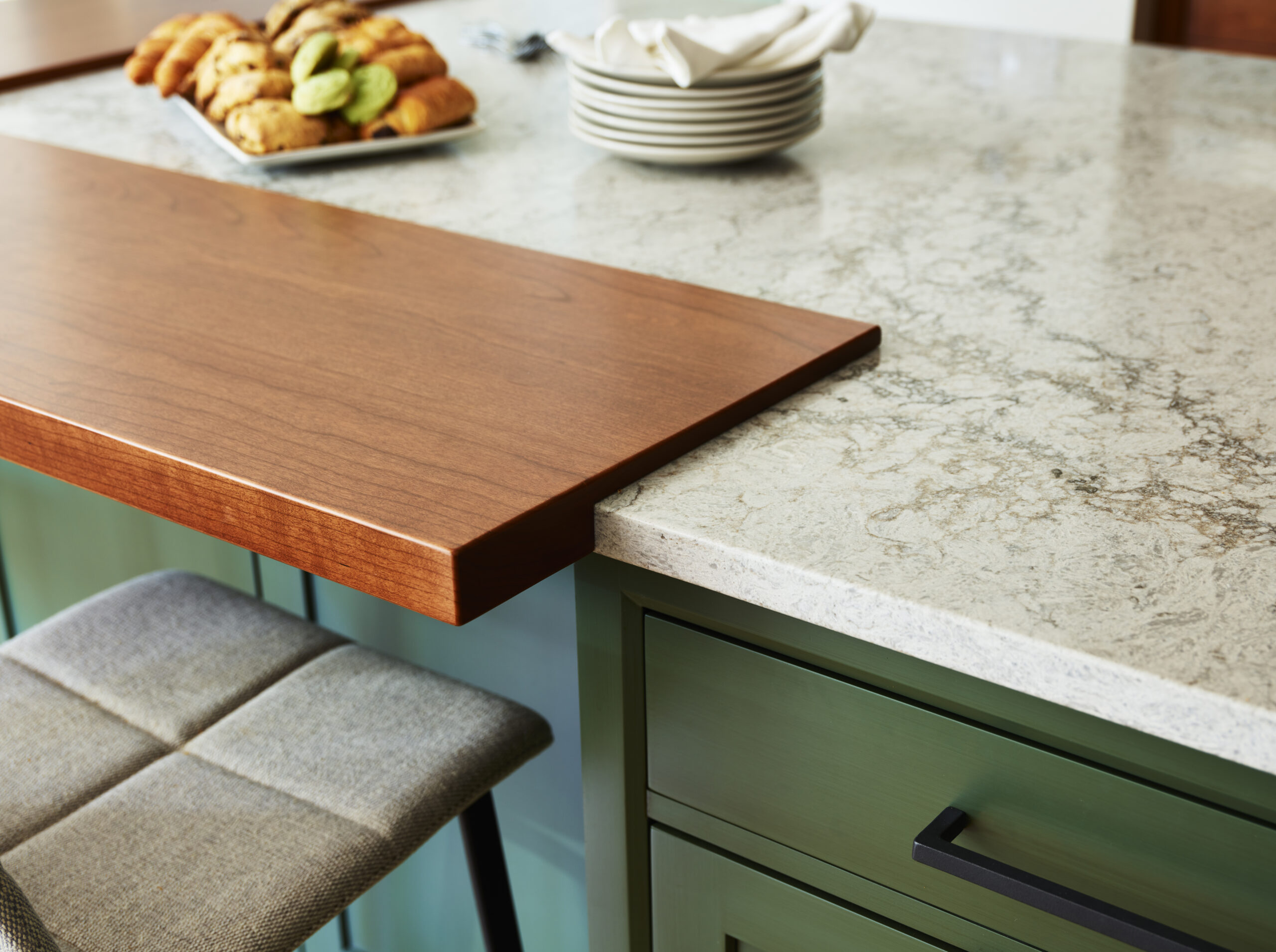 A close-up of a kitchen island with a marble countertop, wooden breakfast bar, green cabinets, and pastries showcases luxury interior design by an experienced interior designer in Massachusetts.