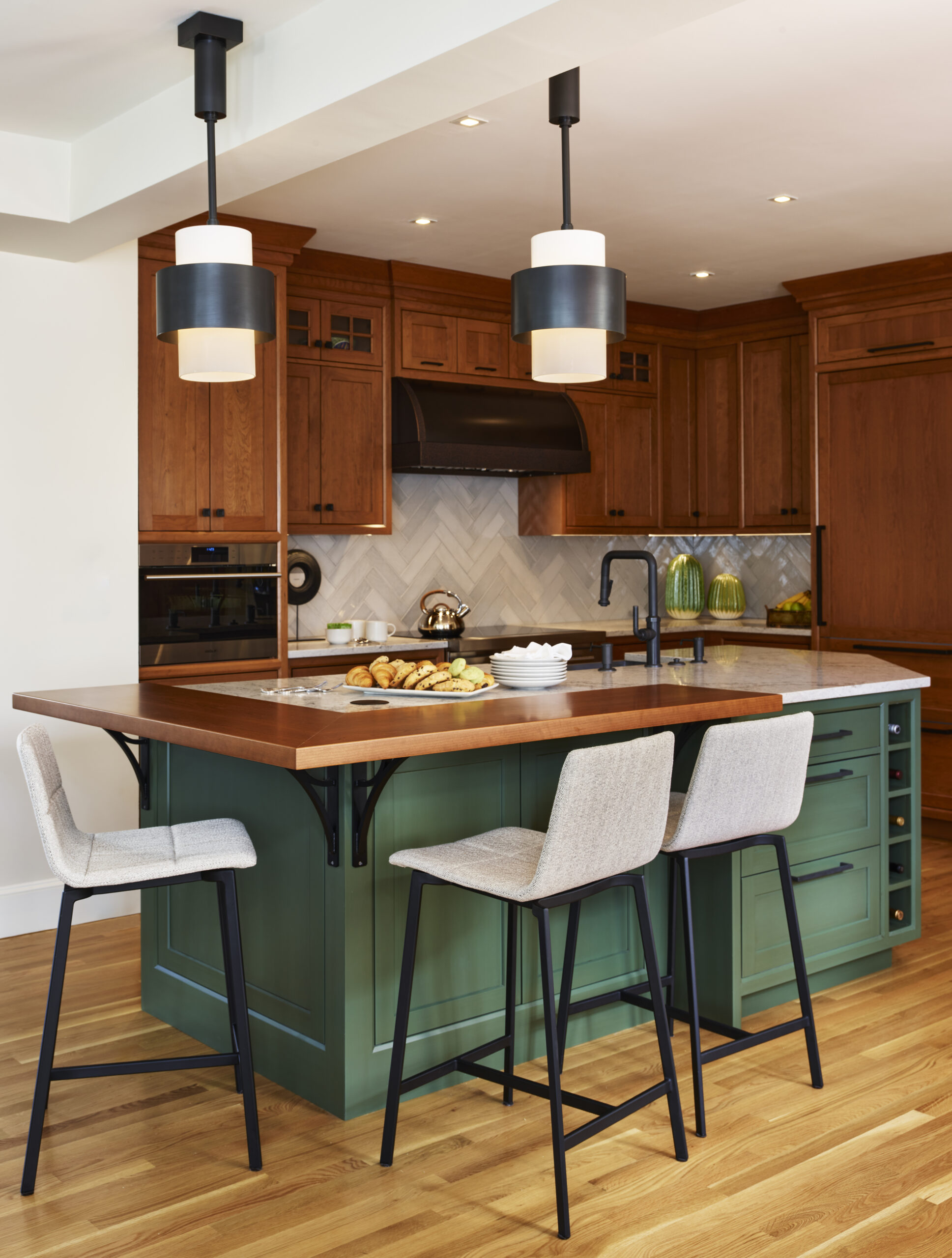 A modern kitchen by an Interior Designer in Massachusetts features wooden cabinets, a green island with a wooden countertop, two upholstered barstools, pendant lights, and baked goods on the counter for a touch of luxury interior design.