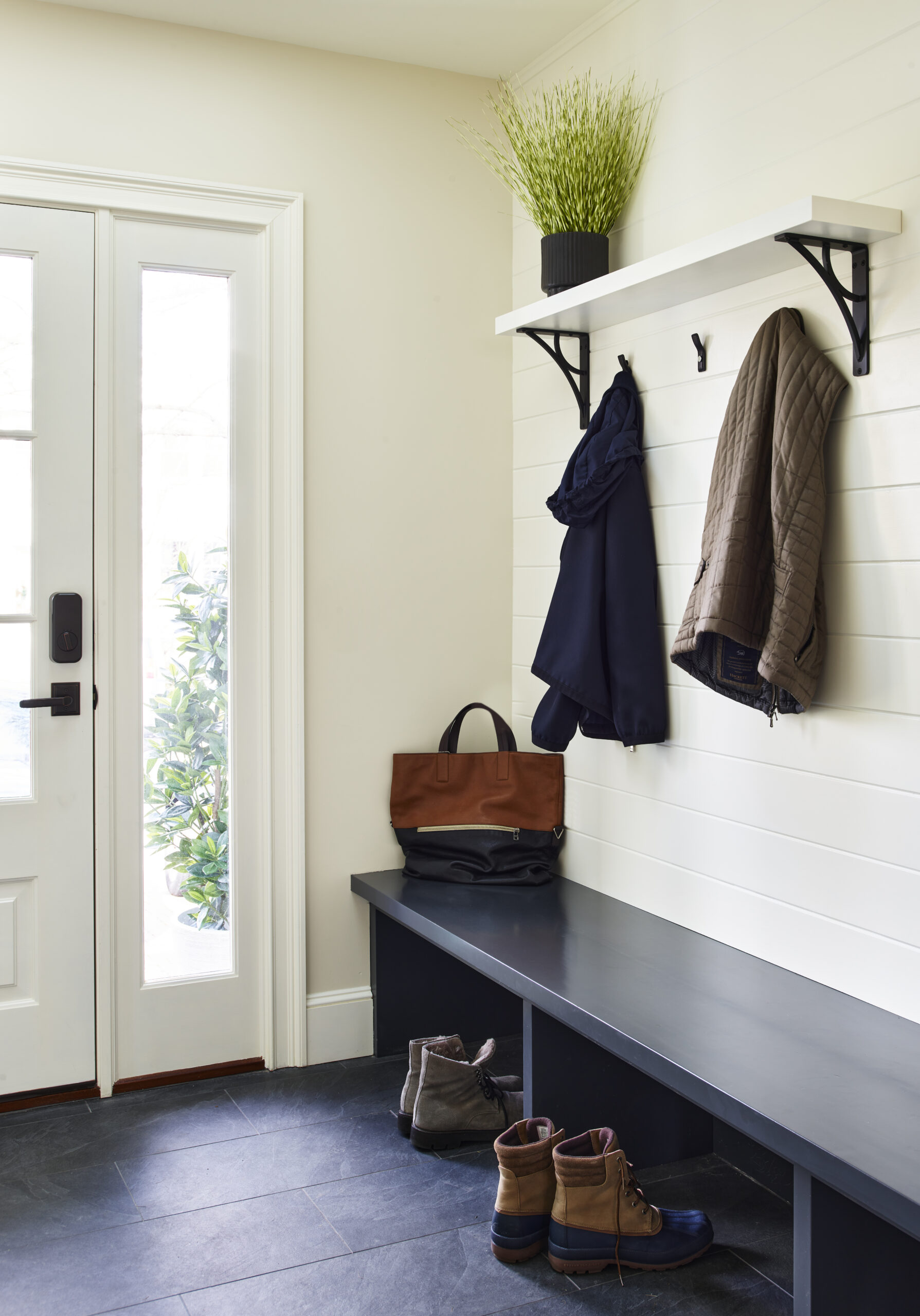 Entryway designed by an interior designer in Massachusetts, featuring a bench, stylish boots, luxury jackets on hooks, a wall shelf with a potted plant, and a glass door bathing the space in natural light.