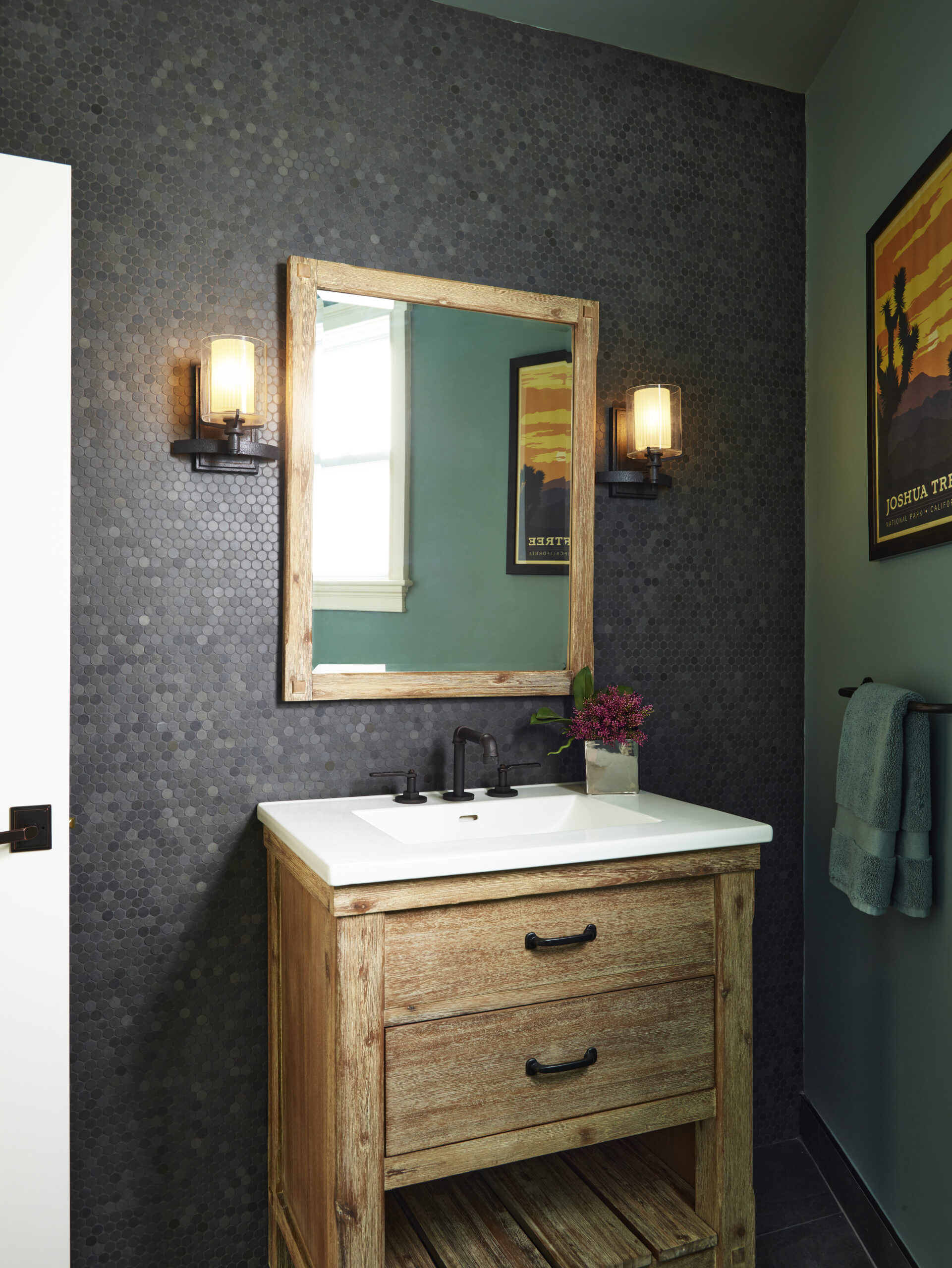 A small bathroom by an Interior Designer in Massachusetts features a wooden vanity with a white sink, a rectangular mirror, wall sconces, dark hex tile backsplash, and a towel hanging by framed art for a touch of luxury interior design.
