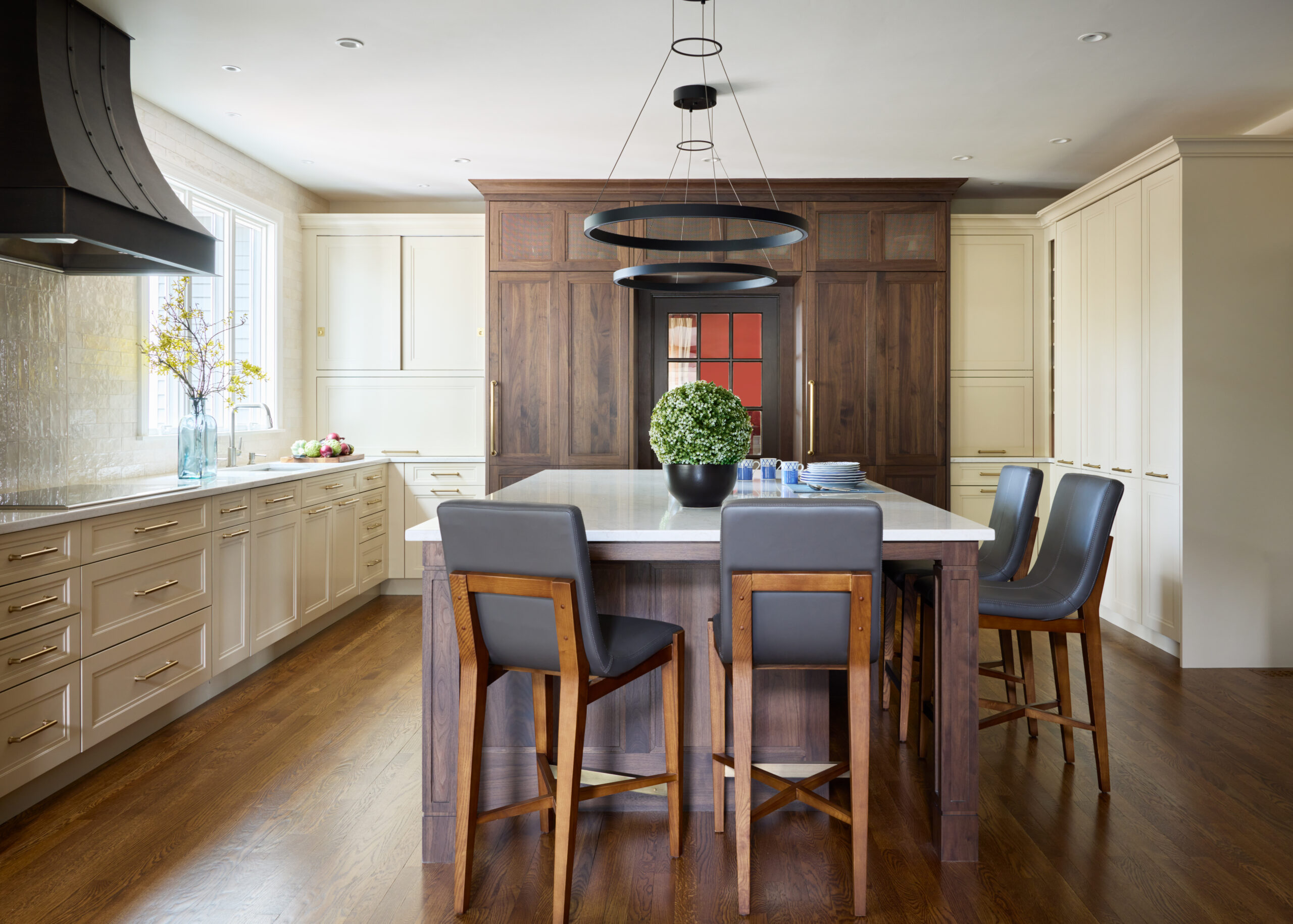 Modern kitchen with a large island, wooden cabinetry, marble countertop, and four chairs surrounds a round plant centerpiece under contemporary pendant lights—a stunning example of luxury interior design by an Interior Designer in Massachusetts.