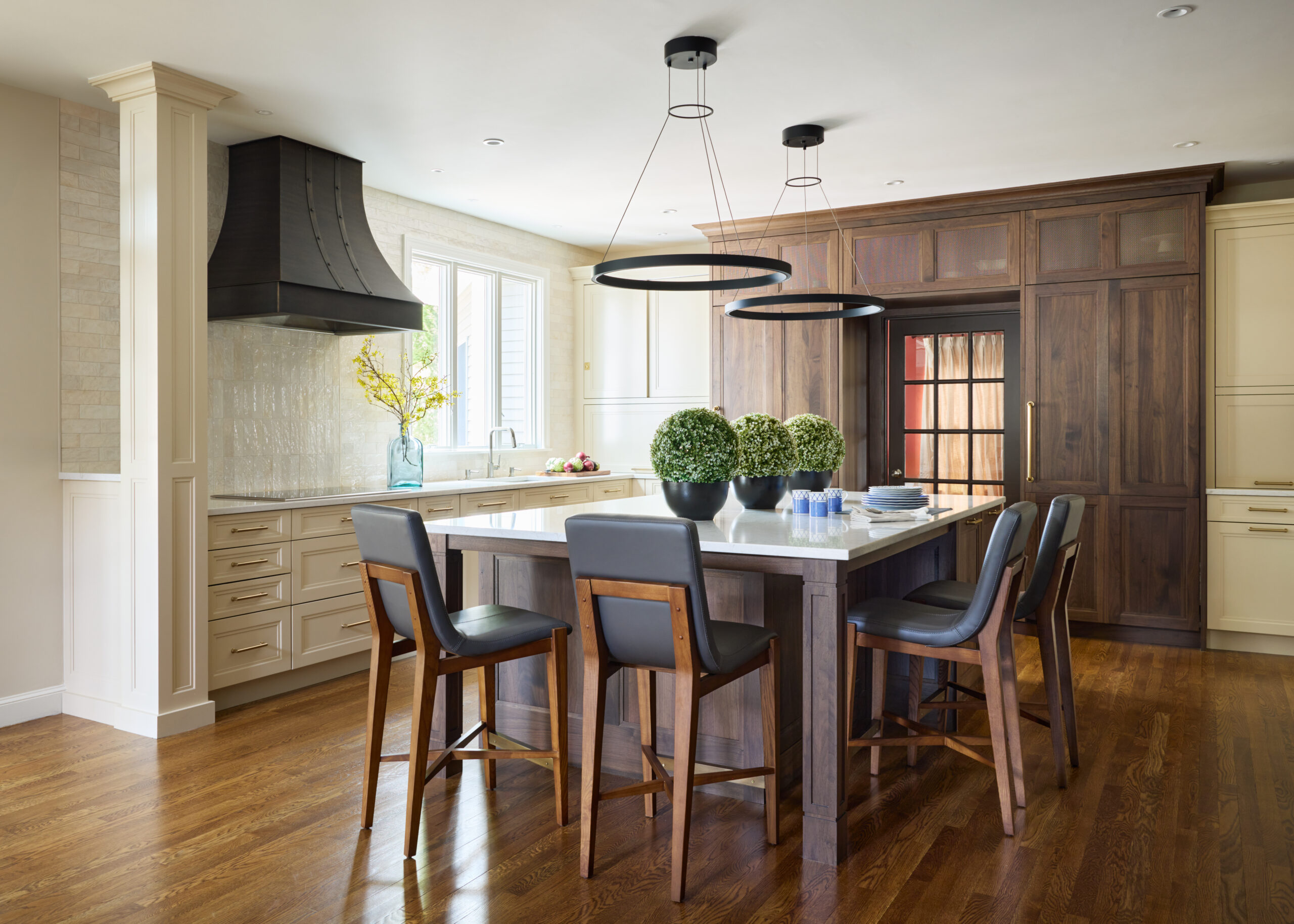 Modern kitchen with a wooden island, four chairs, circular pendant lights, potted plants, wood flooring, and a mix of white and dark wood cabinetry—an example of Luxury Interior Design by top Interior Designers in Massachusetts.
