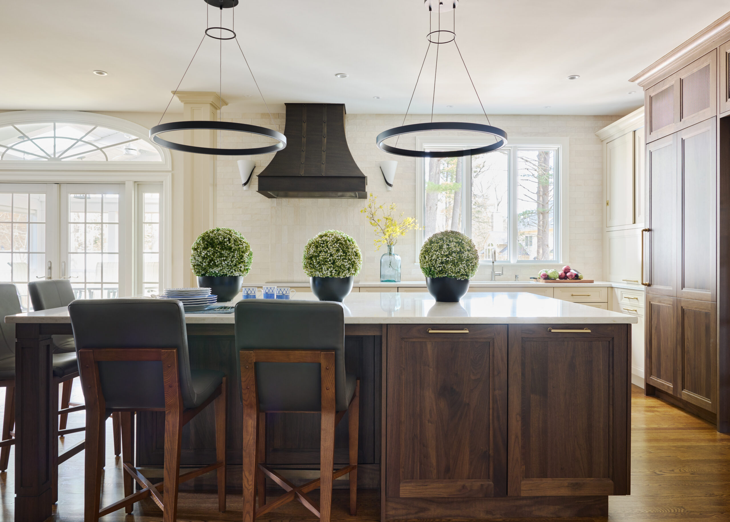 Modern kitchen with wooden cabinets, island with bar stools, two pendant lights, potted plants on the counter, and sunlight streaming through large windows—a stunning example of luxury interior design.