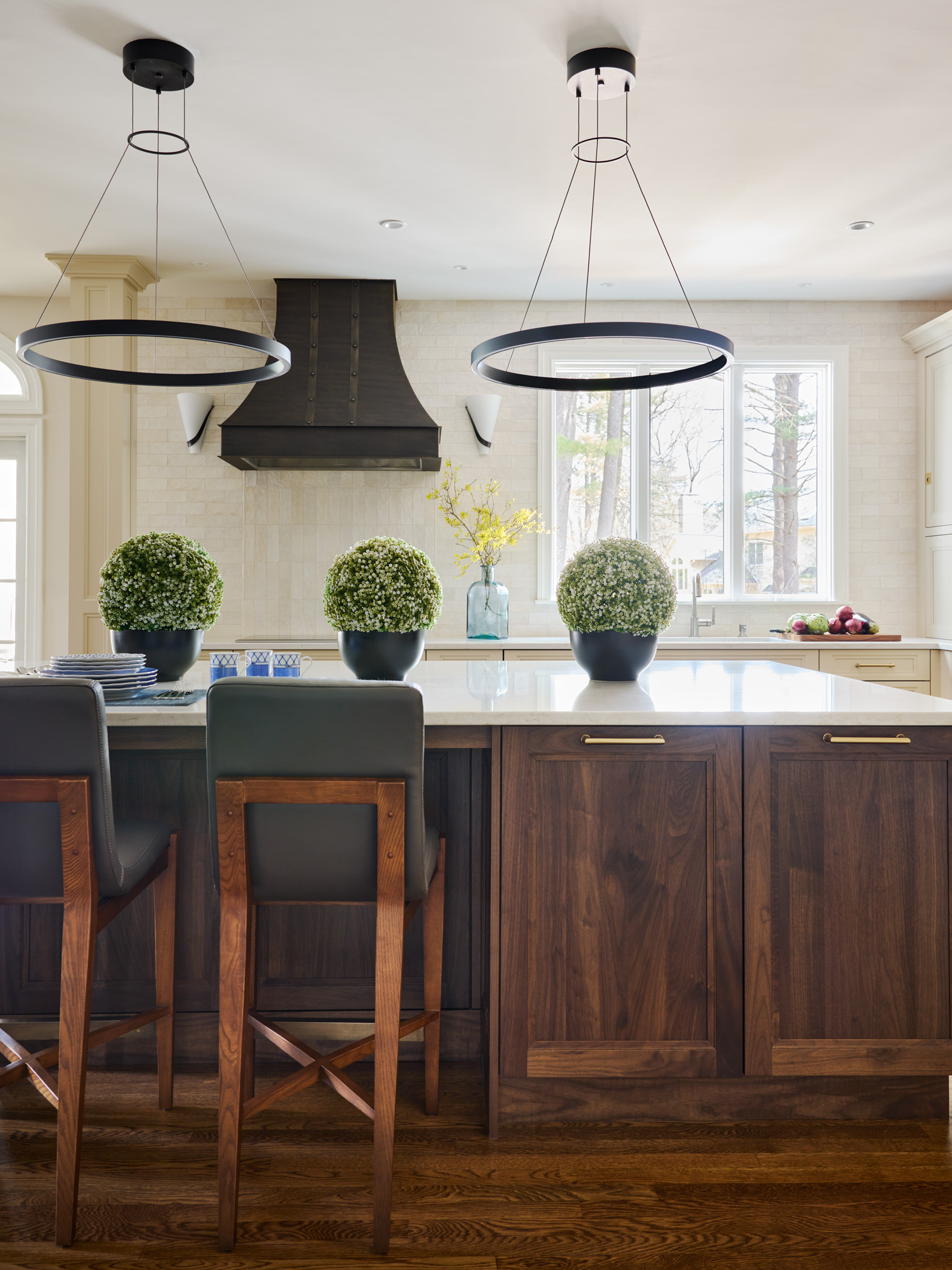 Modern kitchen with dark wood island, luxury interior design touches, two pendant lights, three potted plants on the countertop, and a dark range hood set against a light tile backsplash.