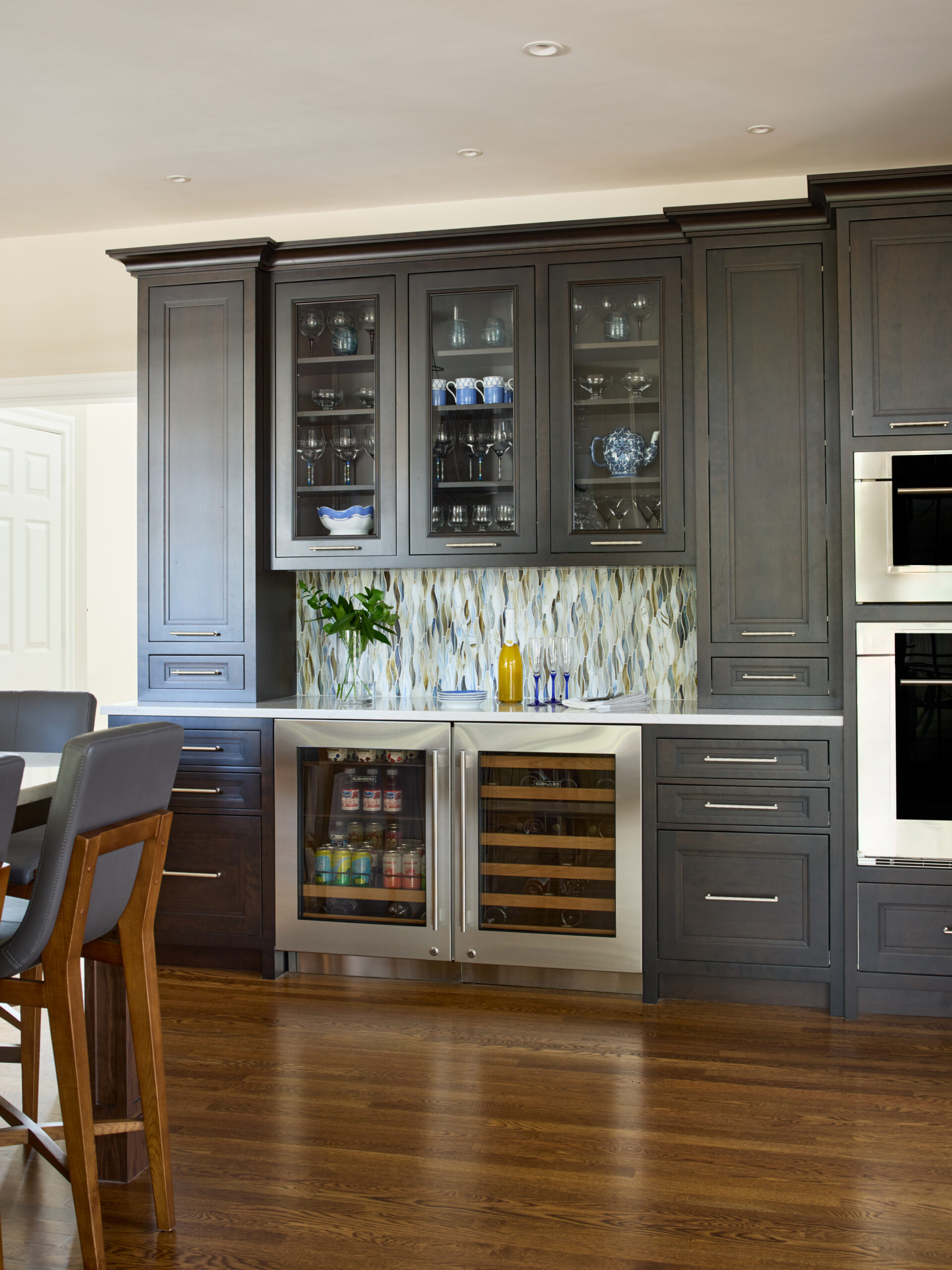 Modern kitchen features dark wood cabinets, a built-in wine fridge, glass-front uppers, and a patterned tile backsplash—an elegant touch of luxury interior design by an Interior Designer Massachusetts. Wood flooring and dining area are partially visible.