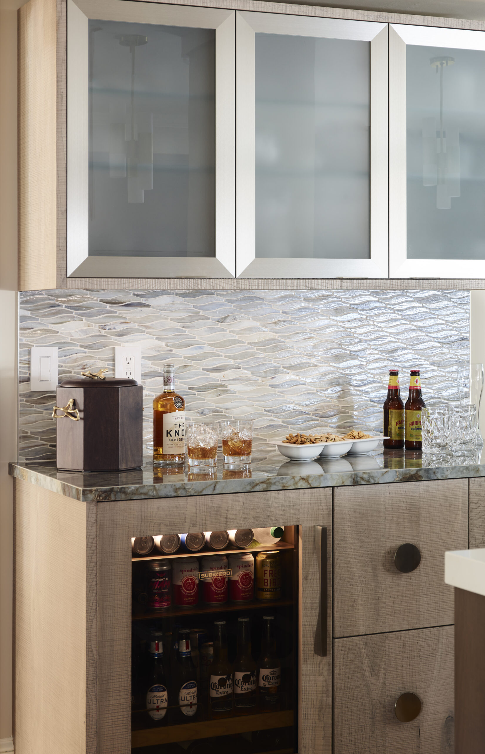 Modern home bar with glass-front cabinets, a small fridge stocked with drinks, bottles of whiskey and beer on the counter, glasses, and a bowl of snacks on a patterned tile backsplash—perfect for a touch of luxury interior design.