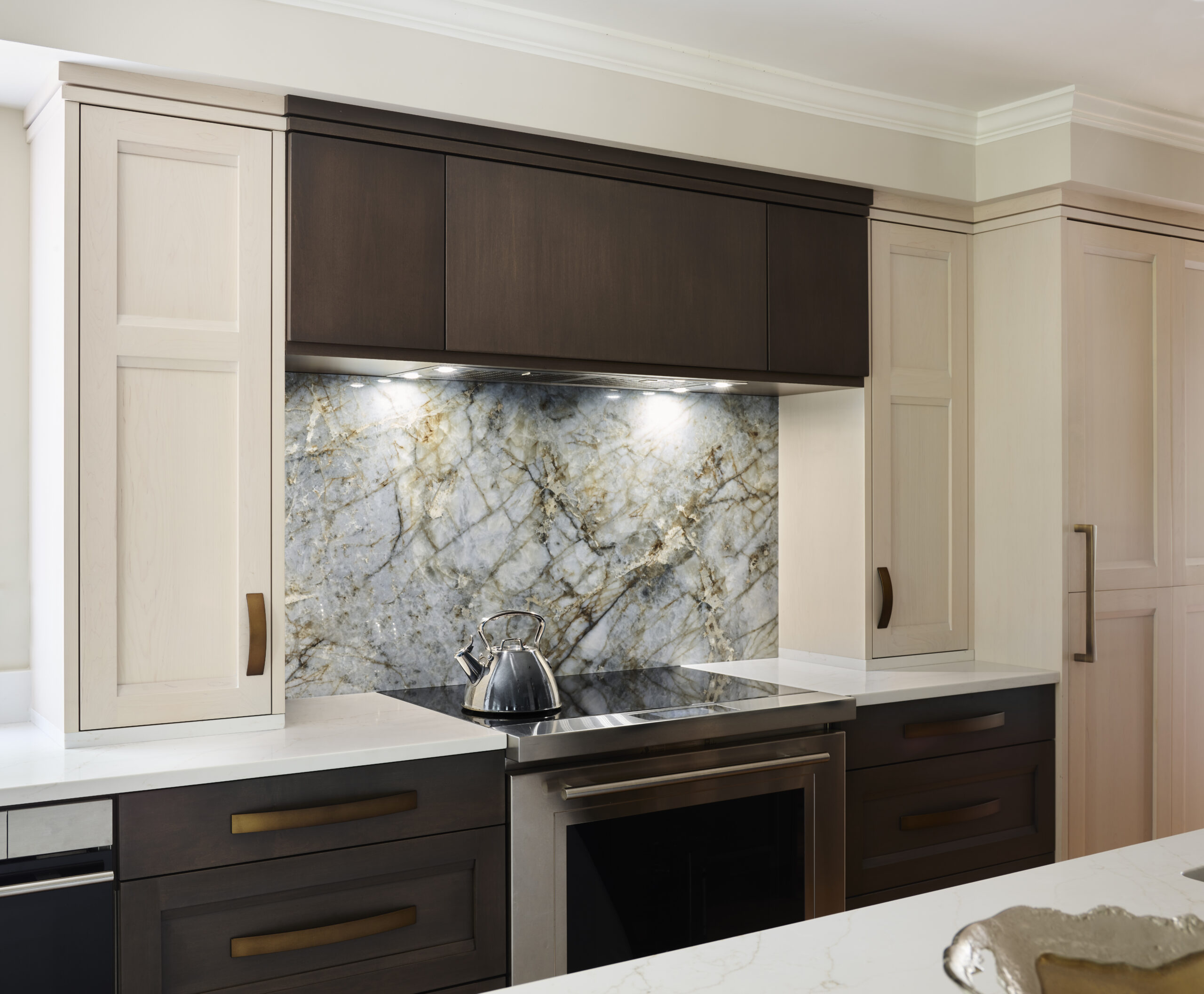 Modern kitchen with dark and light cabinetry, a marble backsplash, an electric stove, and a stainless steel kettle under built-in lights—showcasing luxury interior design by an interior designer in Massachusetts.
