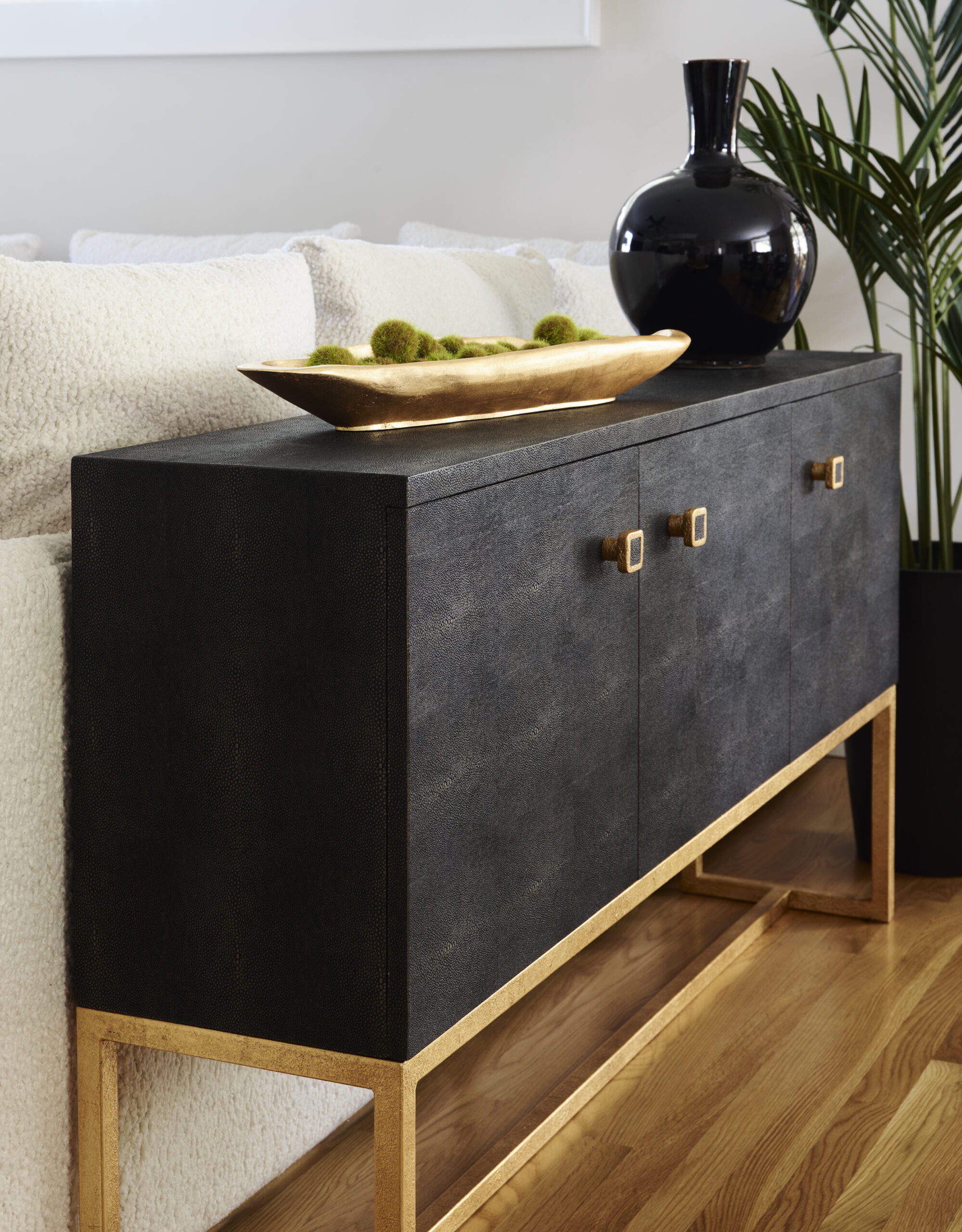 A black and gold sideboard with square handles stands on a wooden floor, topped with a black vase and gold bowl of green spheres, embodying Luxury Interior Design next to a white sofa and tall plant.