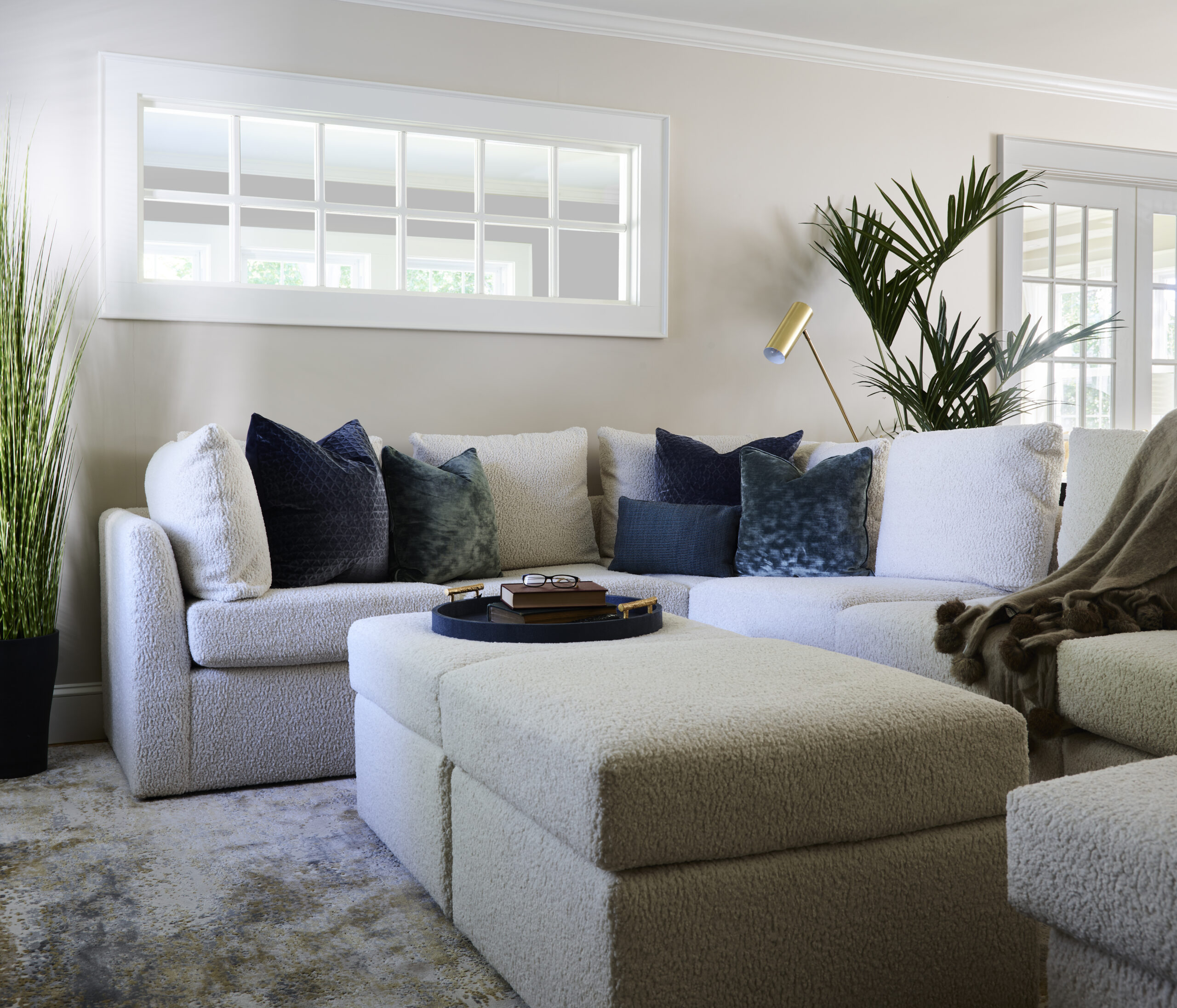 A modern living room curated by an Interior Designer Massachusetts features a white sectional sofa, blue and gray throw pillows, a matching ottoman, gold lamp, and potted plants beside a window with square panes.