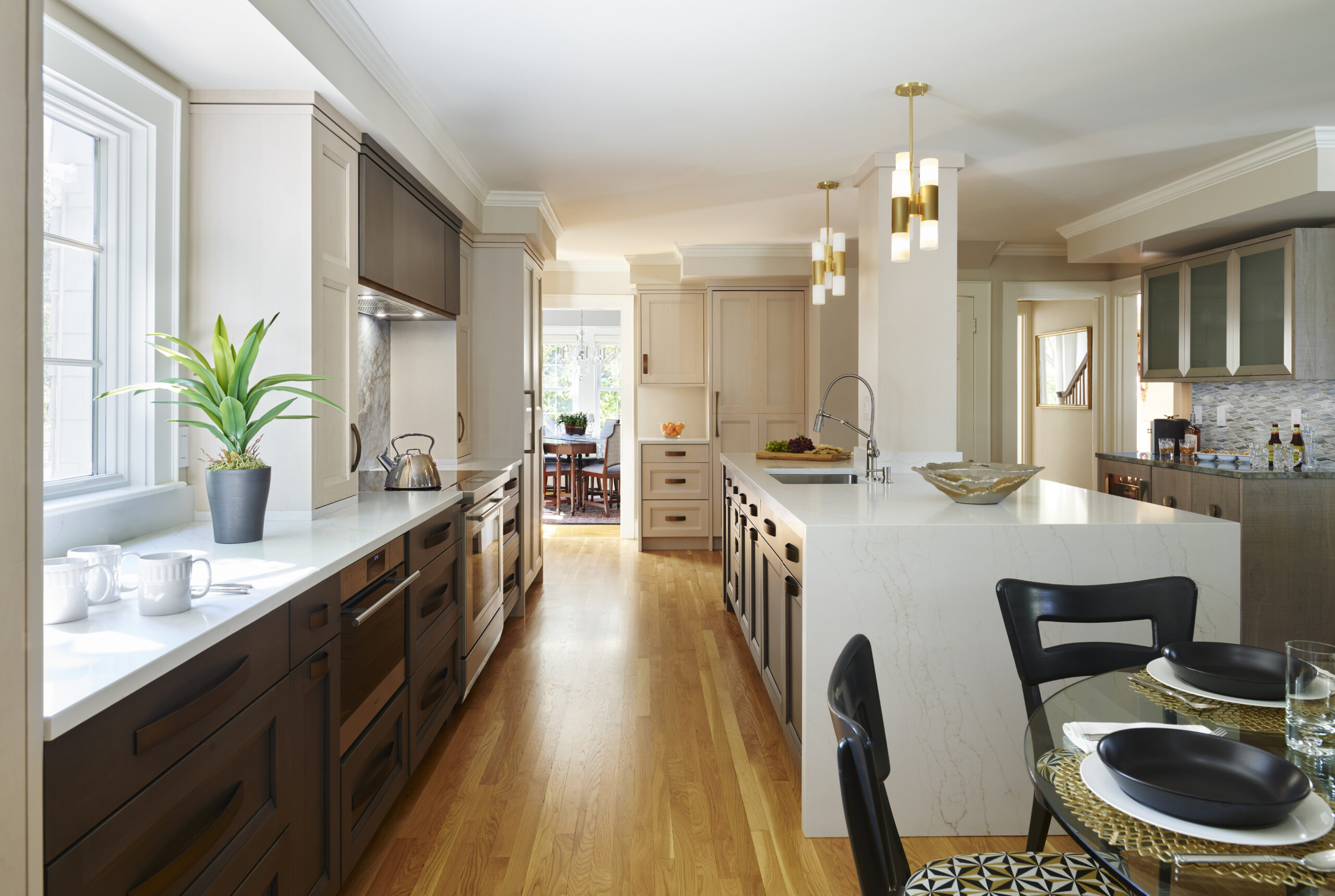 Modern kitchen with wood flooring, white countertops, dark and light cabinetry, a central island with sink, potted plant by the window, and a dining area in the background—a beautiful example of Luxury Interior Design by an Interior Designer Massachusetts.