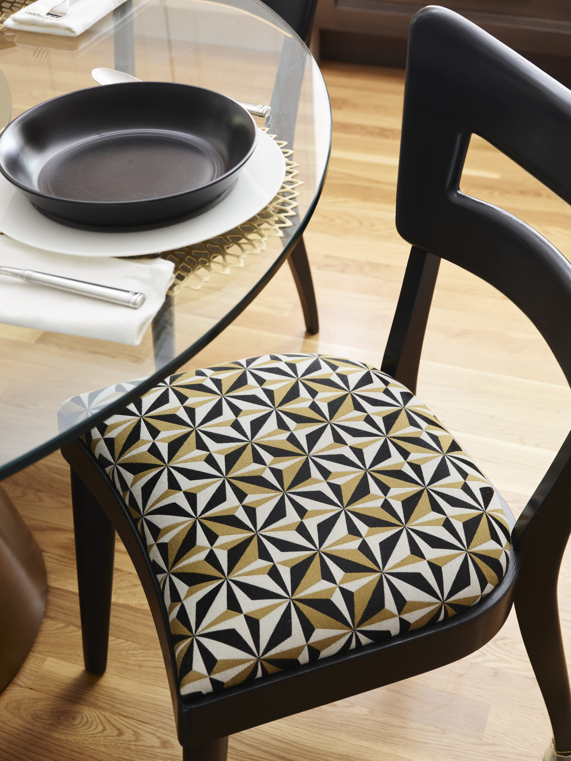 A black wooden chair with a geometric-patterned cushion is positioned next to a glass dining table set with elegant touches—a black bowl, white plate, napkin, and gold placemat—reflecting the sophistication of luxury interior design.